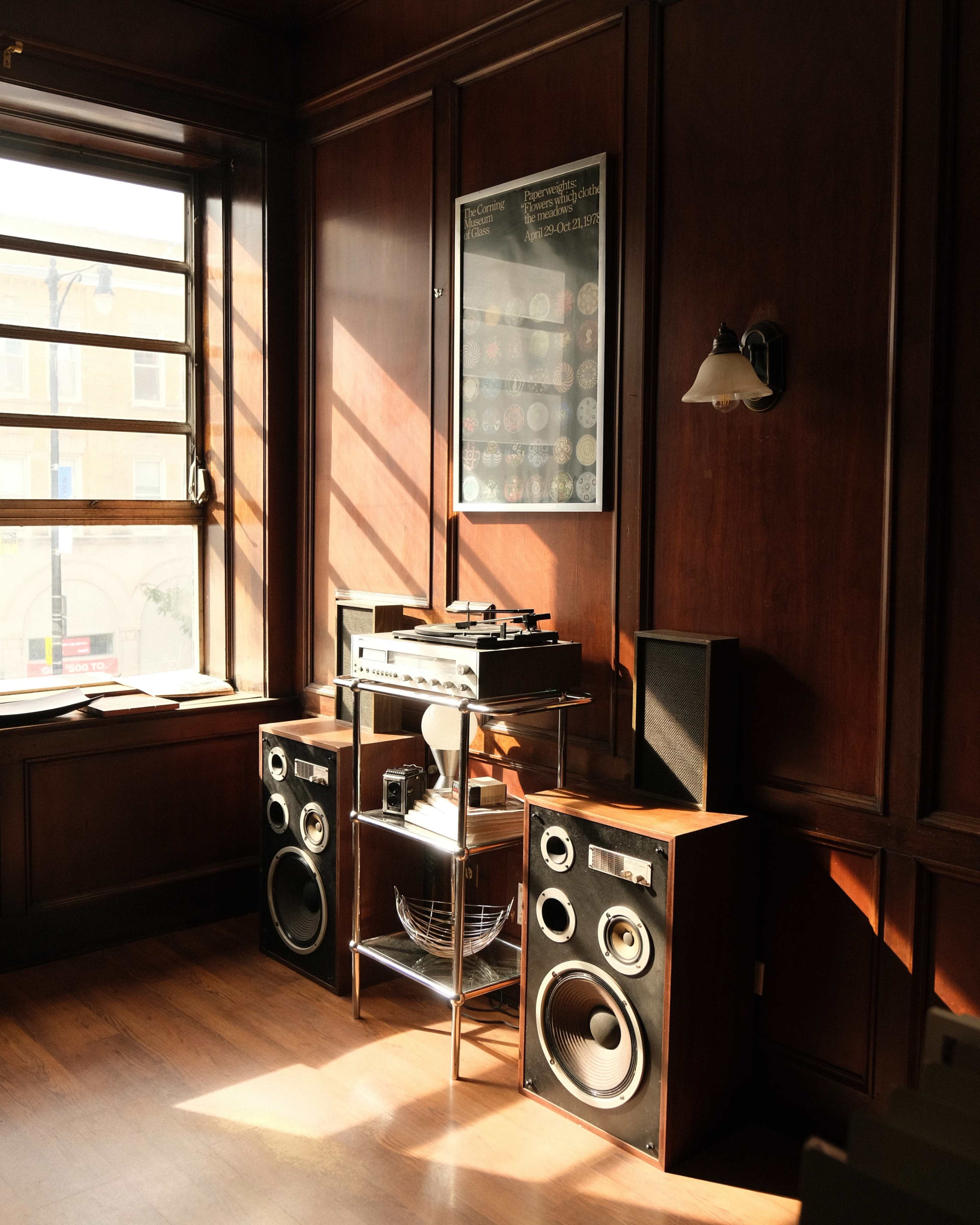 A wooden-walled room features a record player and large speakers beside a window, illuminated by sunlight.