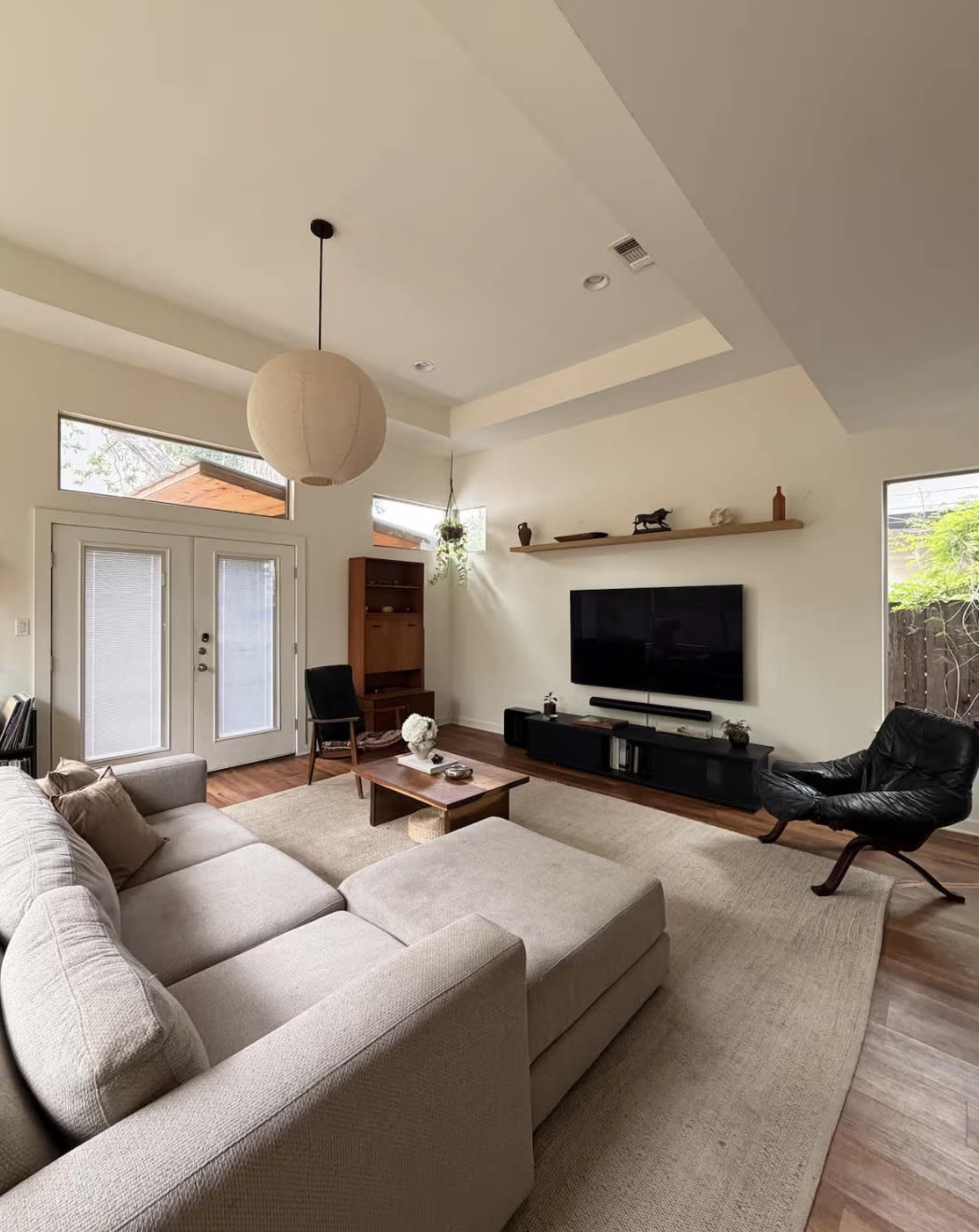 A modern living room features a light-colored sectional sofa, a wooden coffee table, a television mounted on the wall, and large windows allowing natural light.