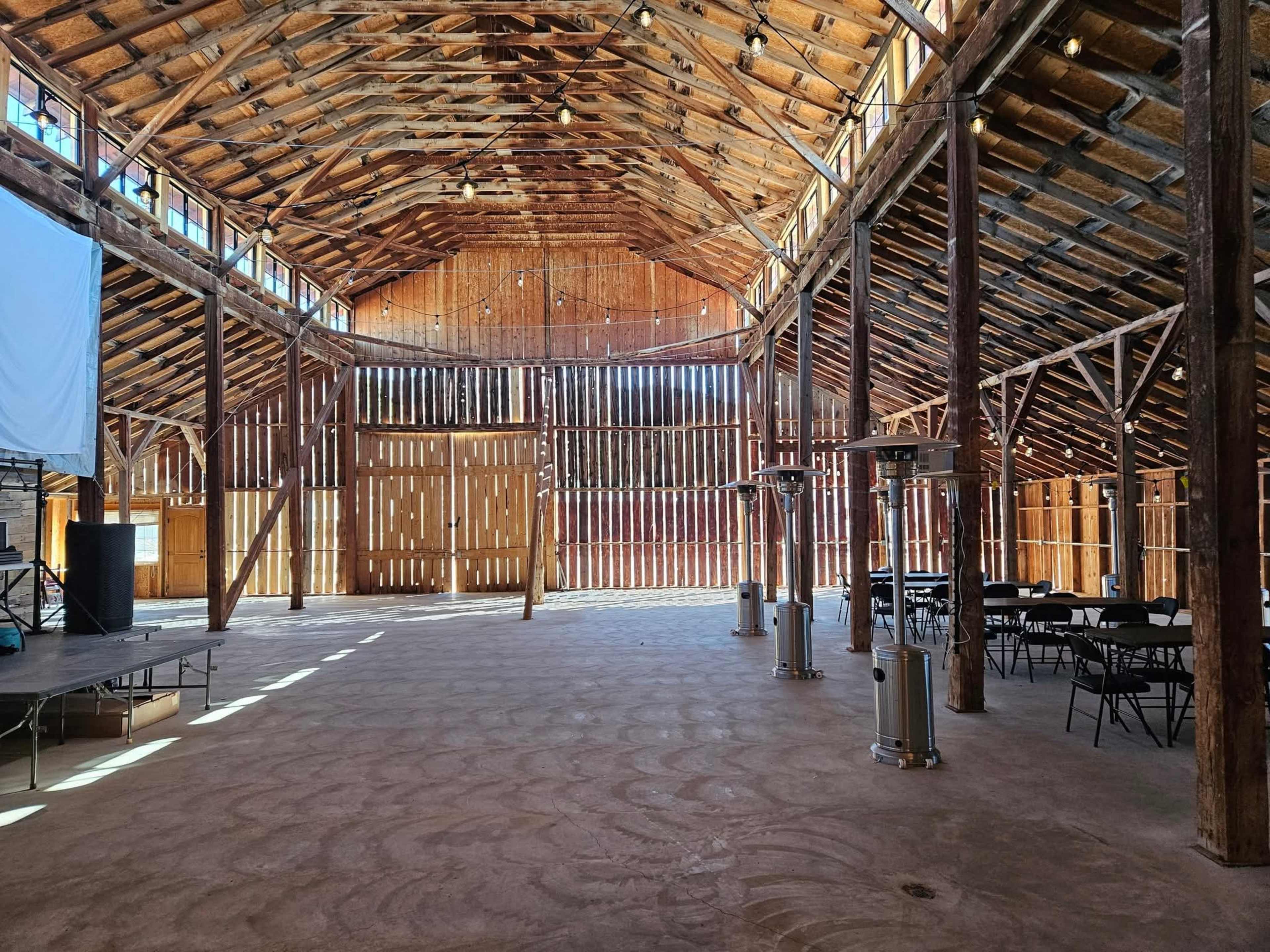 The interior of a spacious wooden barn features high ceilings, exposed beams, and scattered chairs with heating lamps in the corners.