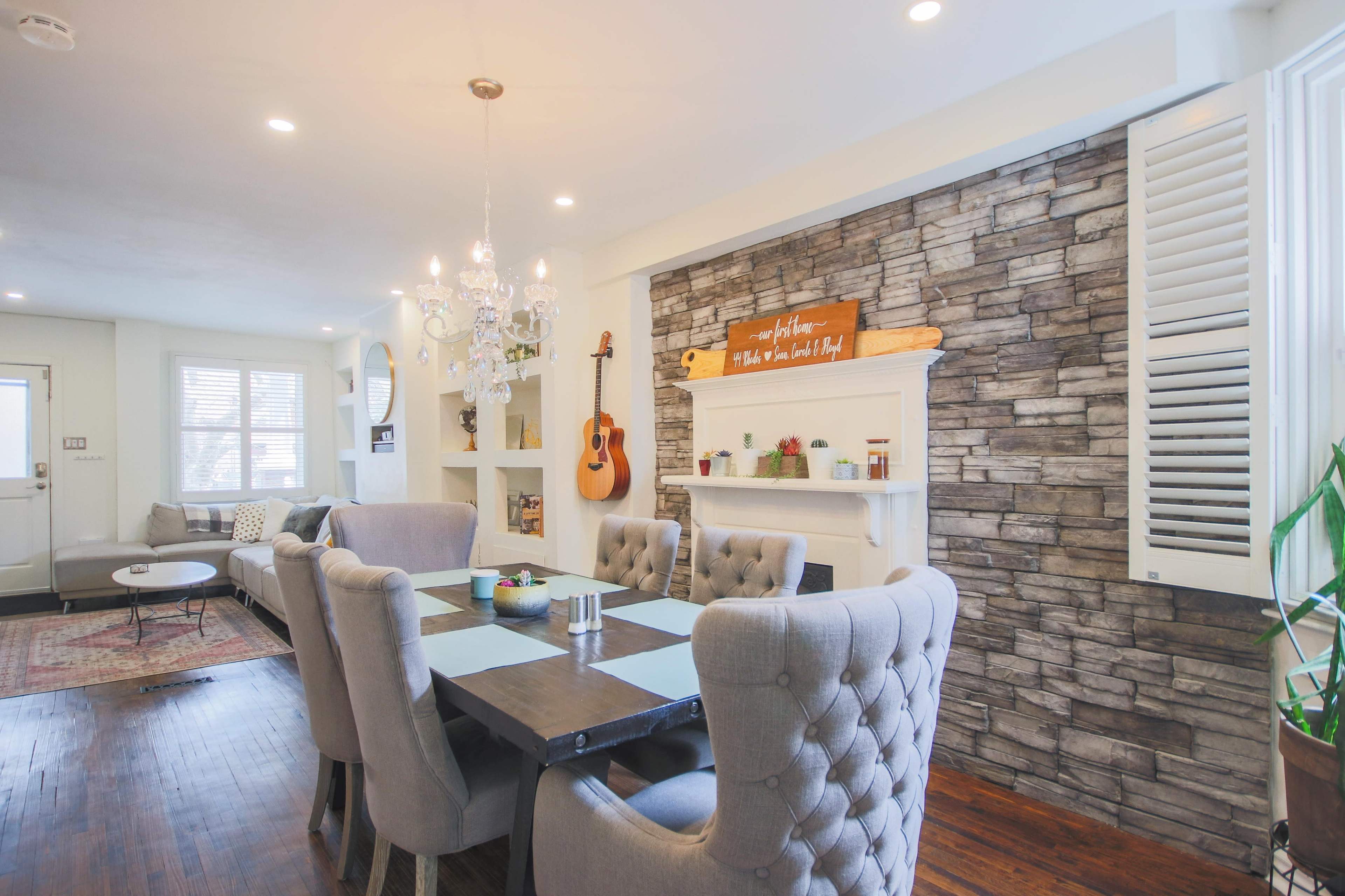 The image shows a dining area with a wooden table surrounded by upholstered chairs, a stone accent wall, and decorative elements including a chandelier and a guitar.