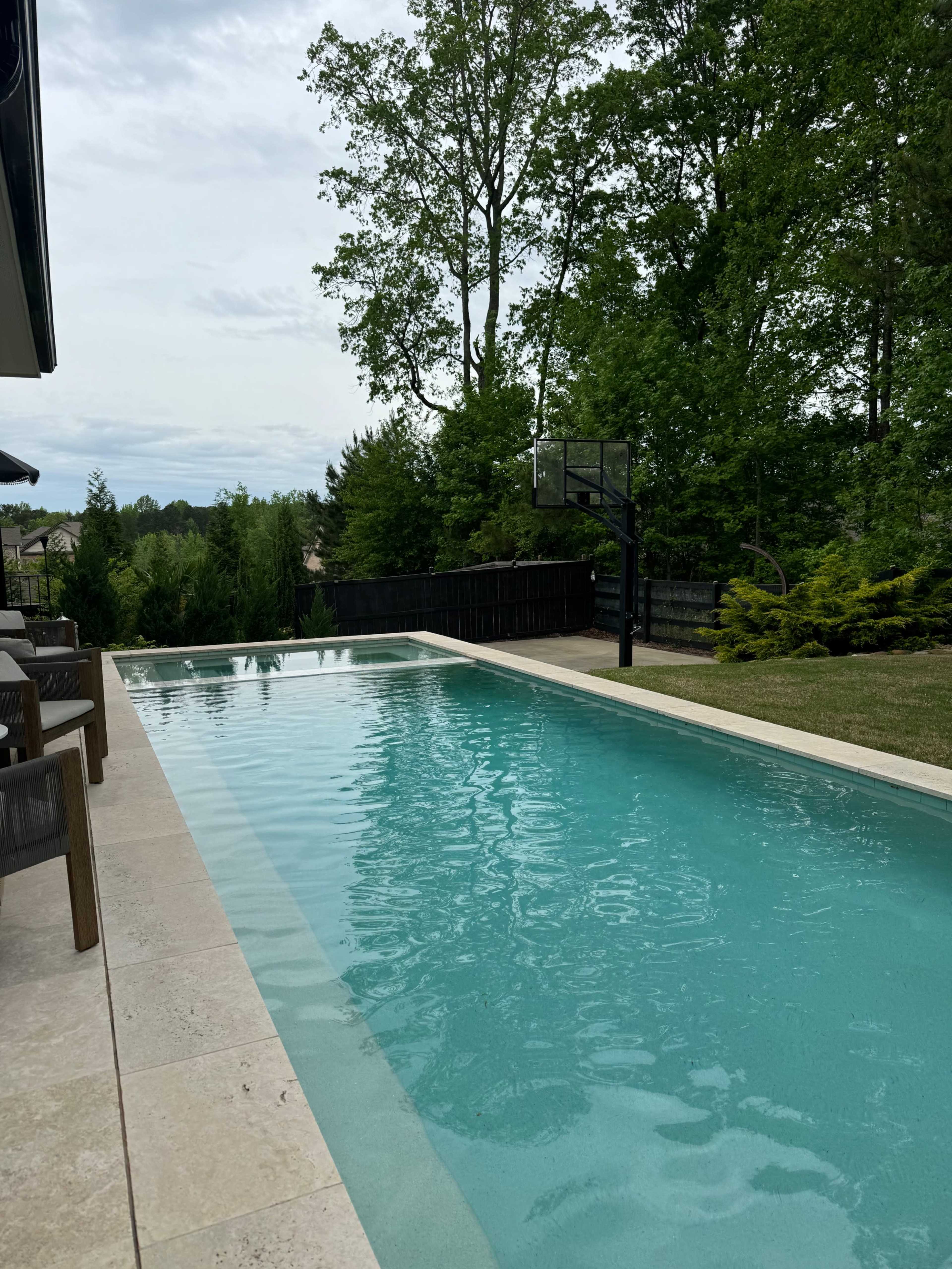 A swimming pool with clear water is set beside a grassy area and a basketball hoop, surrounded by trees and a wooden fence.