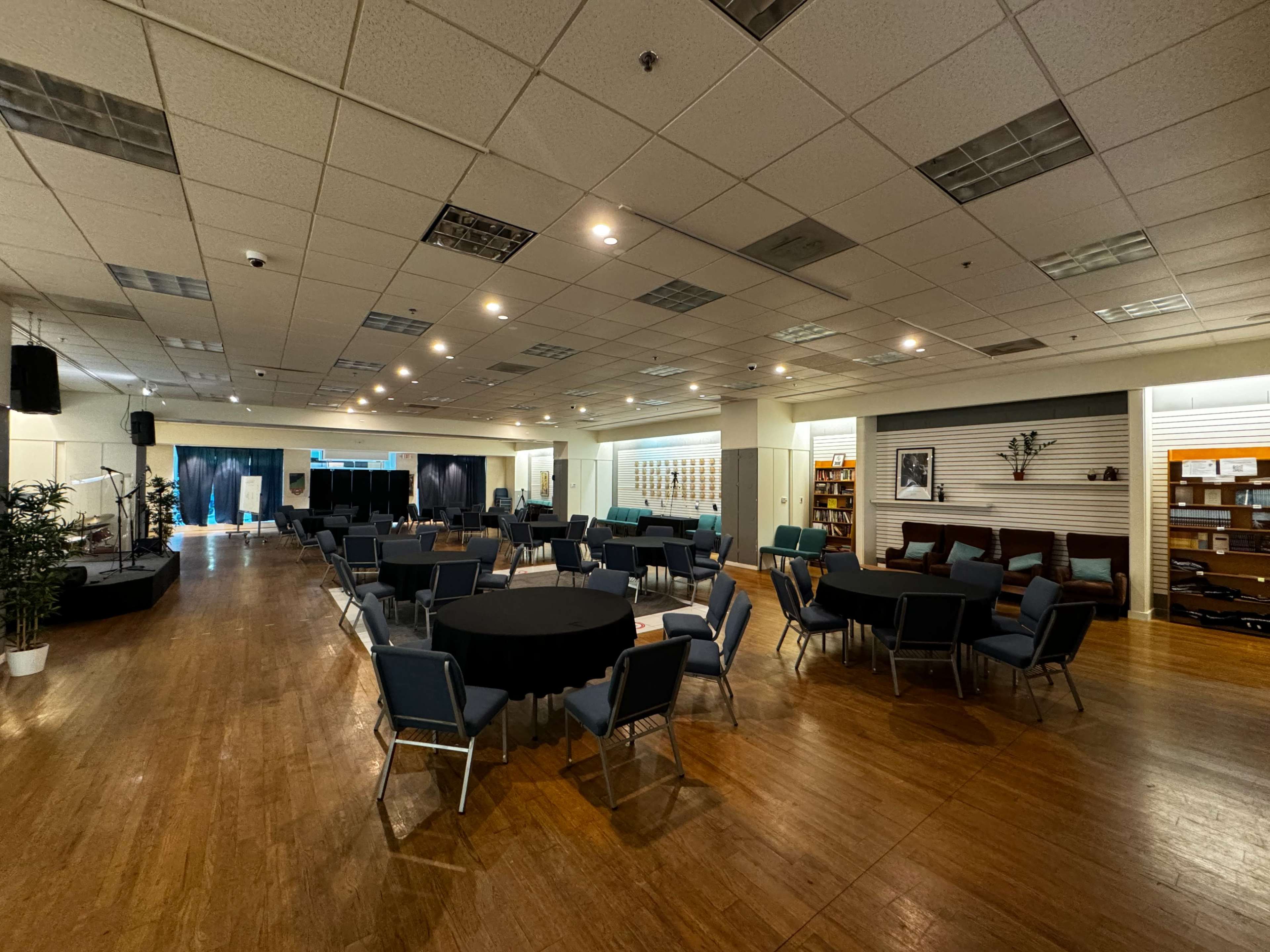 The image shows a spacious indoor event area with round and square tables arranged, along with a stage on one side and shelves in the background.