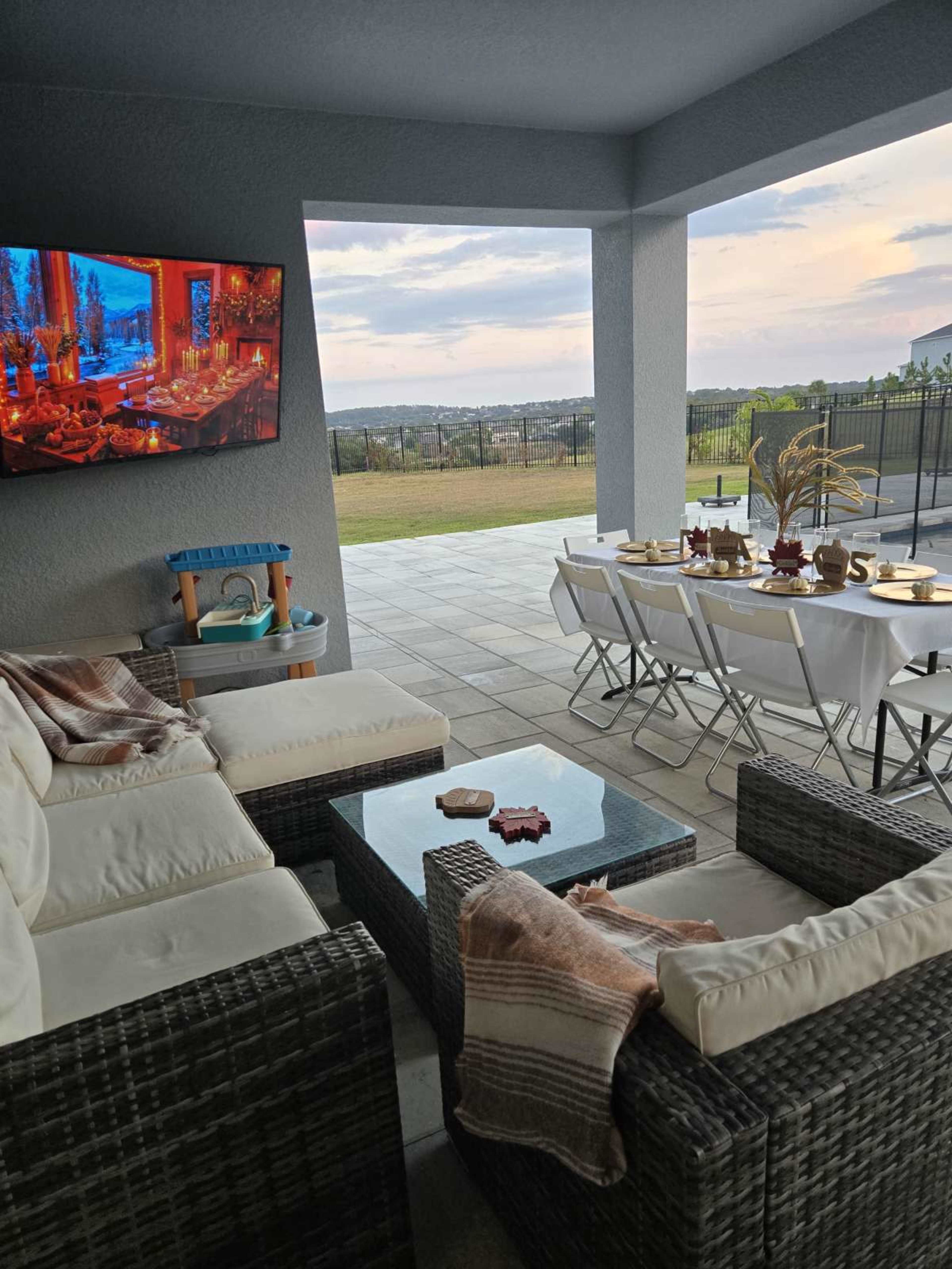 A covered patio features a seating area with a couch and coffee table, a dining setup with chairs and a table, and a television mounted on the wall.