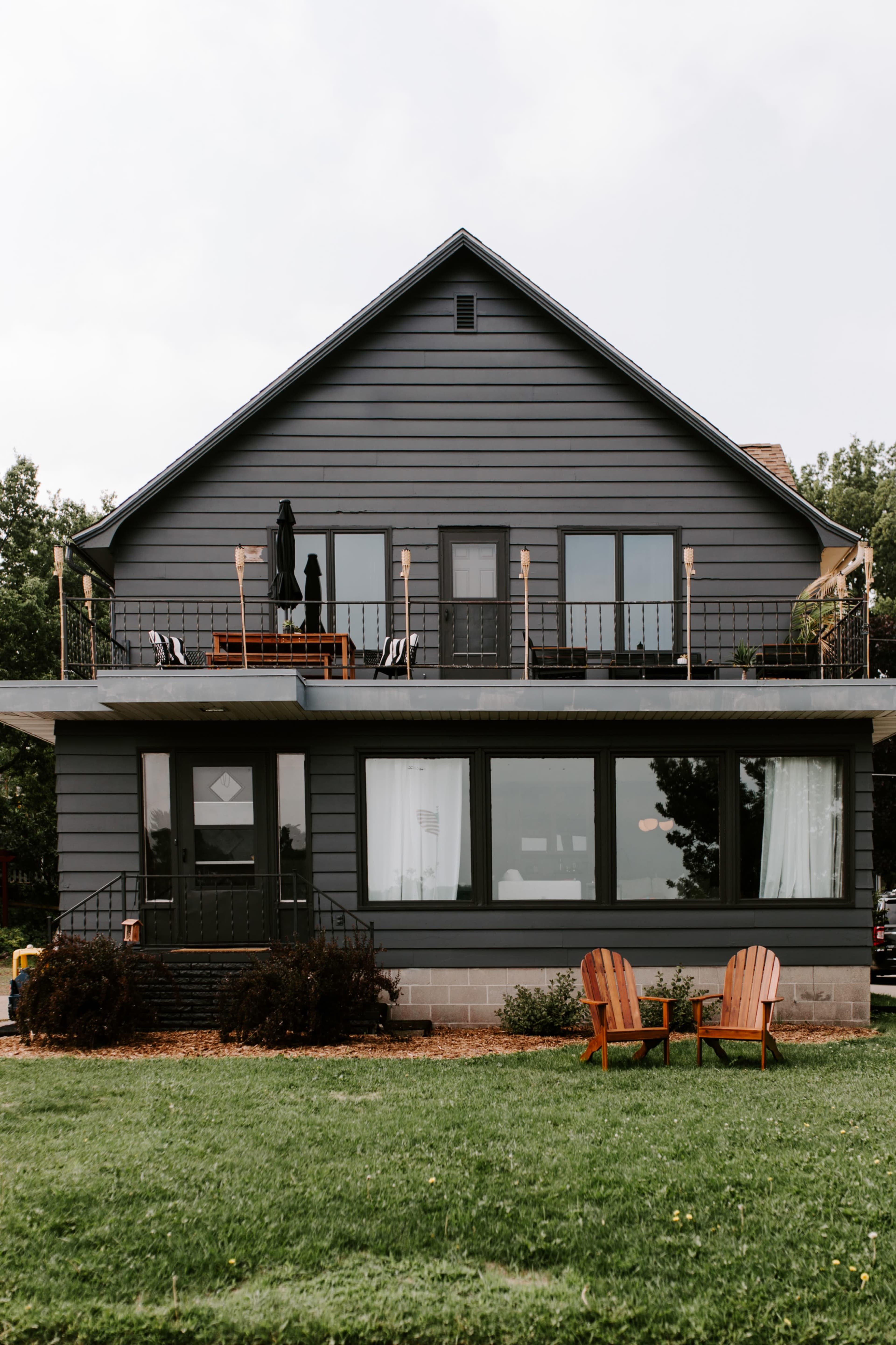 A gray two-story house features a front porch with seating and two wooden chairs on a grassy lawn.
