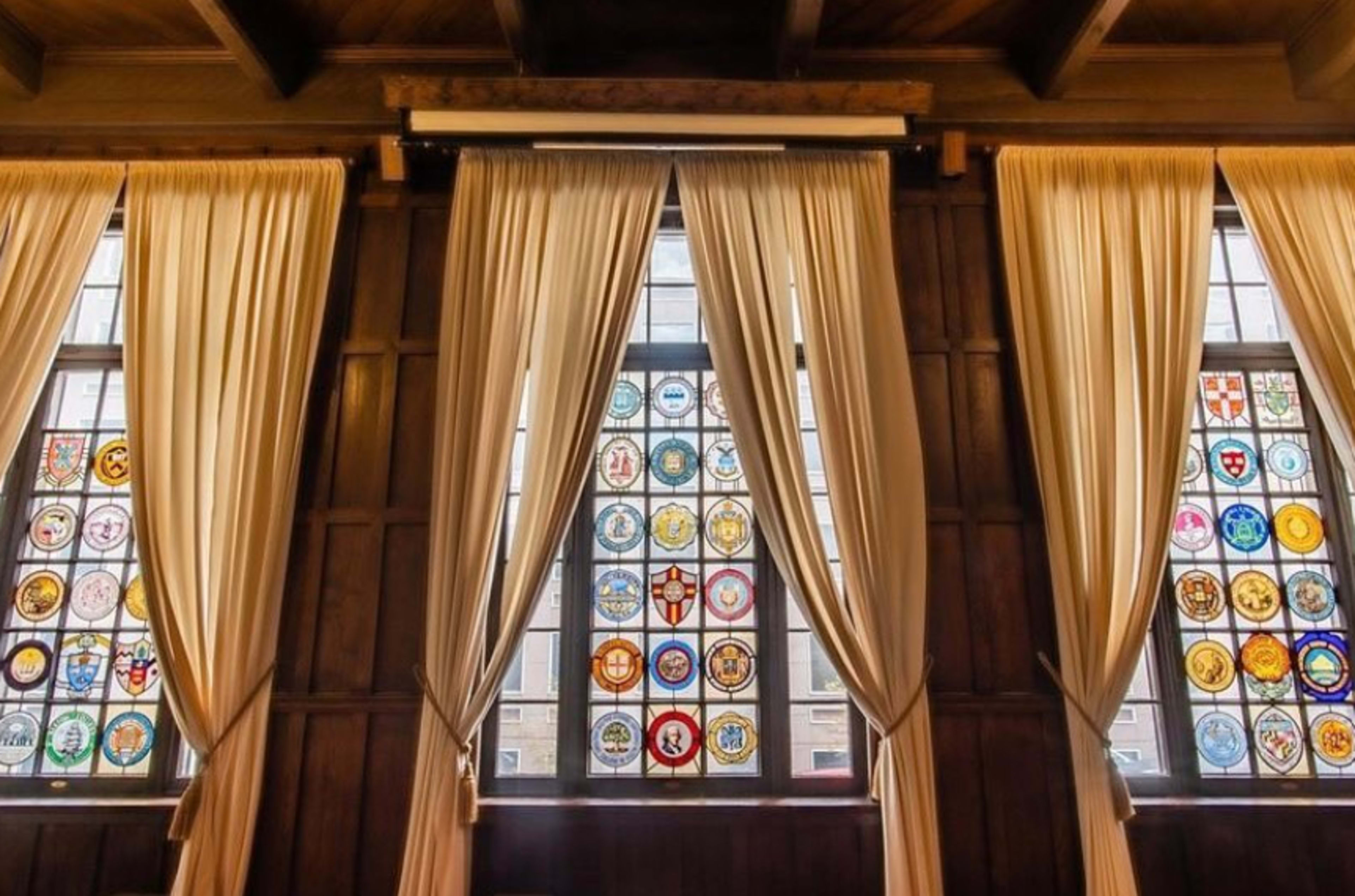Three tall windows featuring stained glass panels with various emblems are framed by cream-colored curtains in a wooden-paneled room.