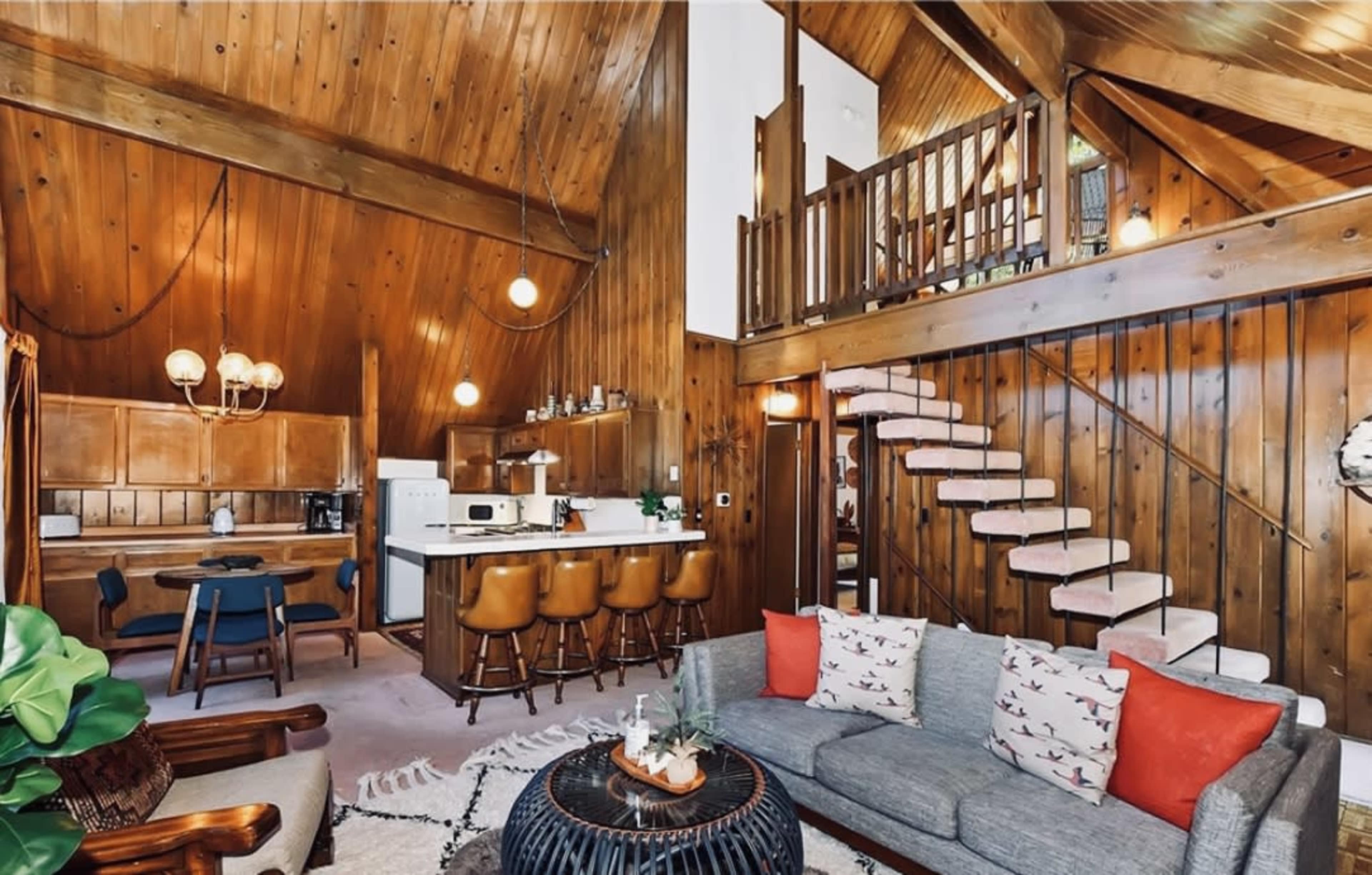 A wooden-paneled living space featuring a staircase, a gray sofa, a dining area, and a kitchen.