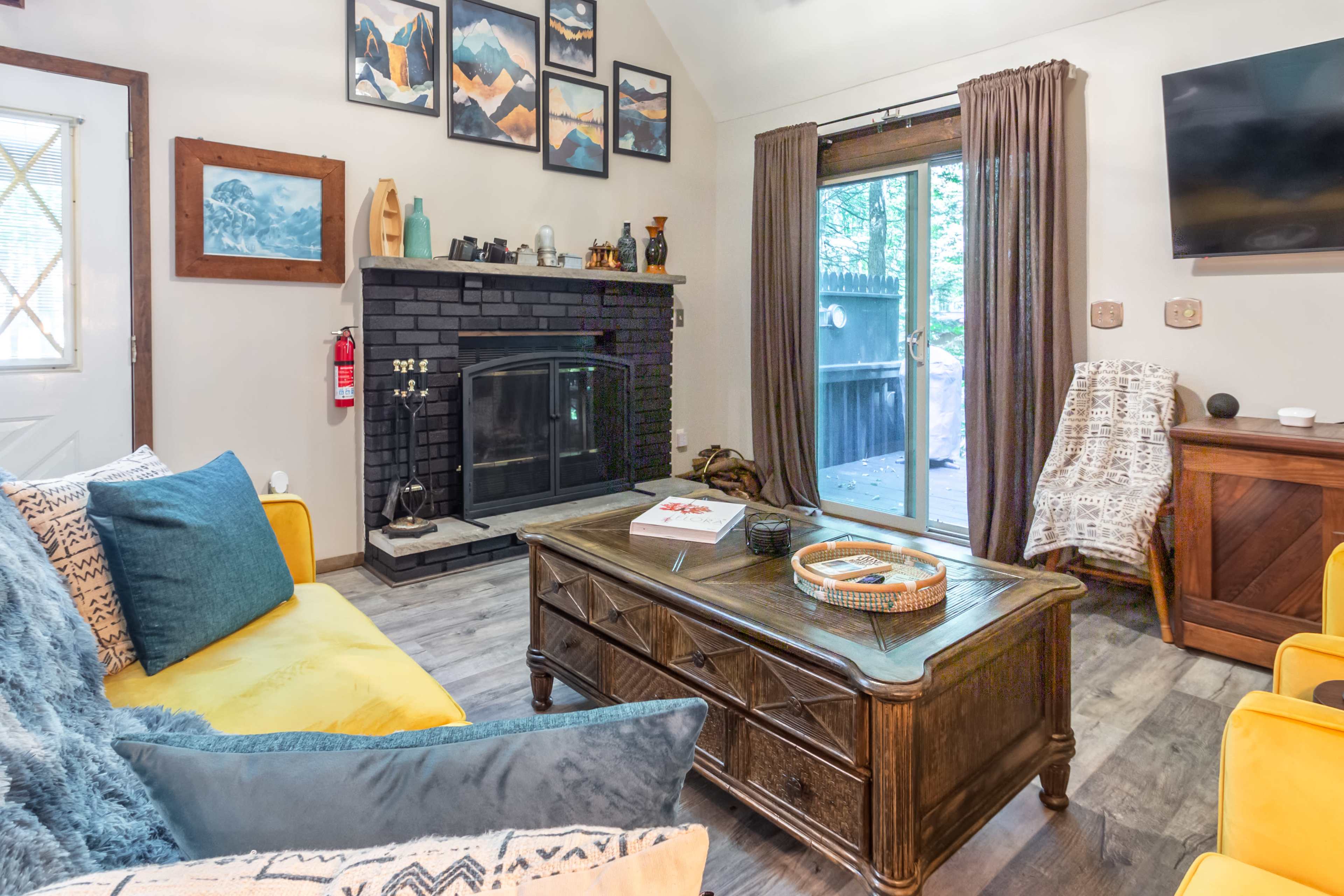 A cozy living room features a yellow sofa, a wooden coffee table, a black fireplace, and large windows opening to a deck.