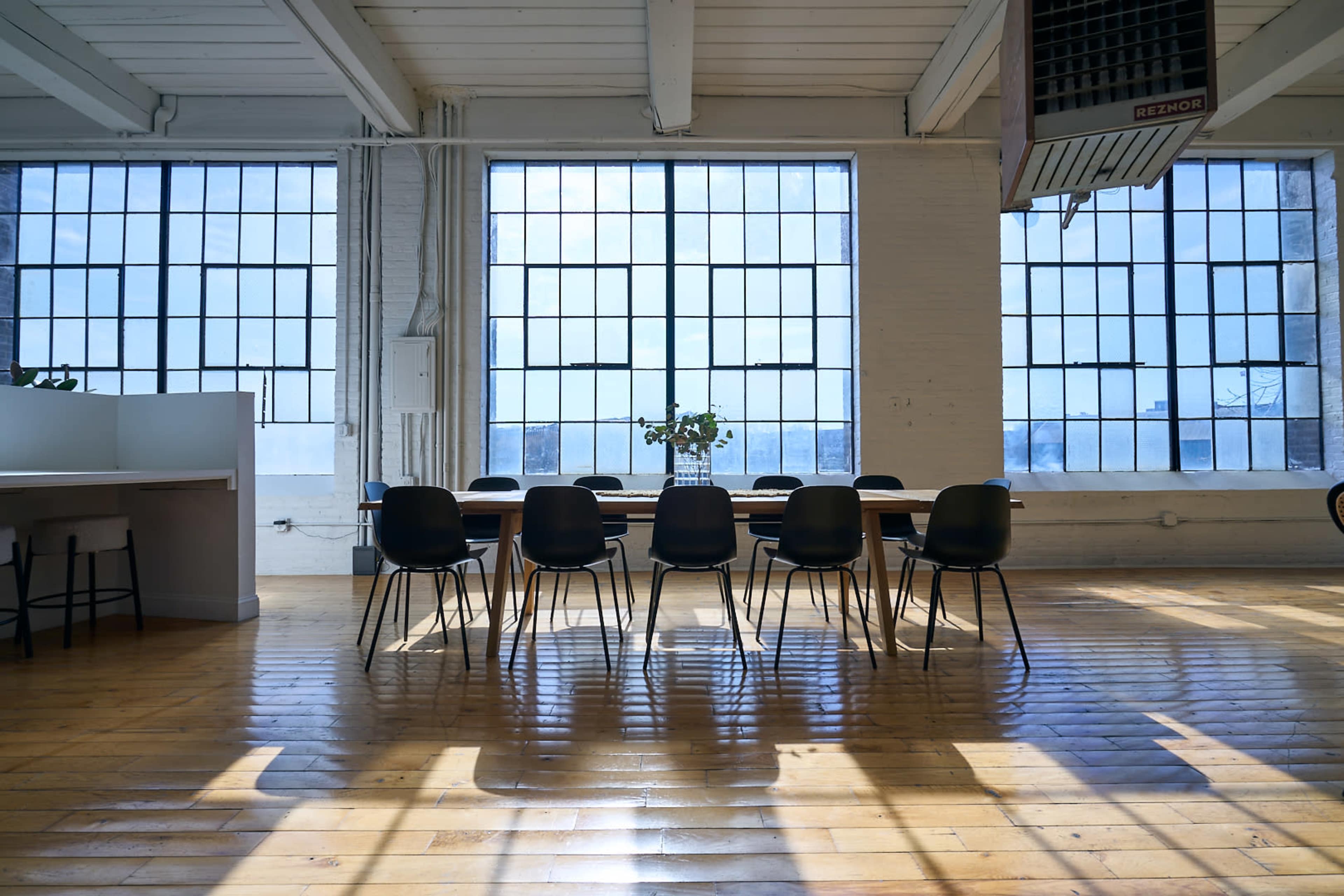 A long table surrounded by chairs is positioned in an airy room with large windows that let in natural light.