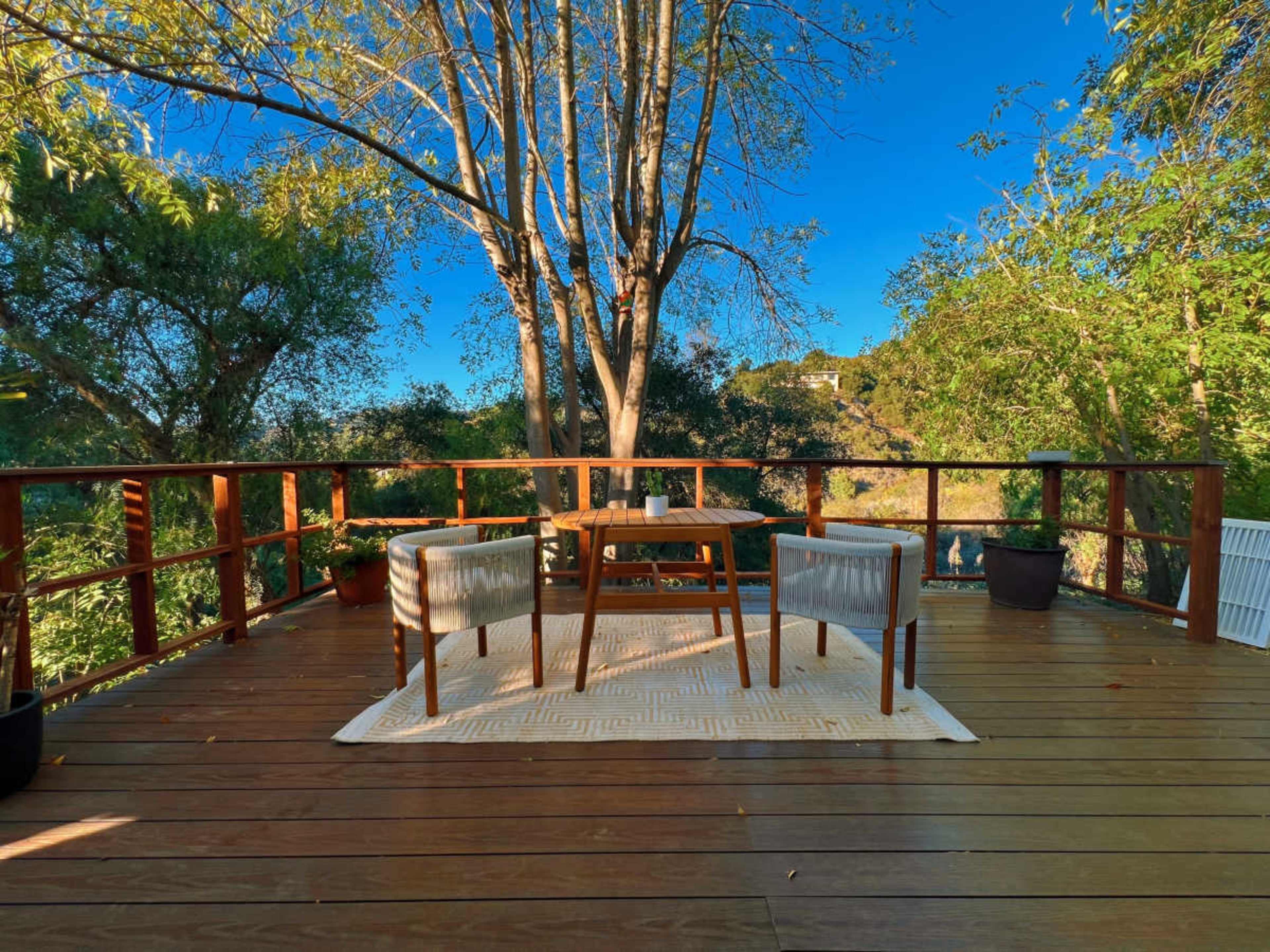 A wooden deck features a table and two chairs, surrounded by trees and greenery under a clear blue sky.