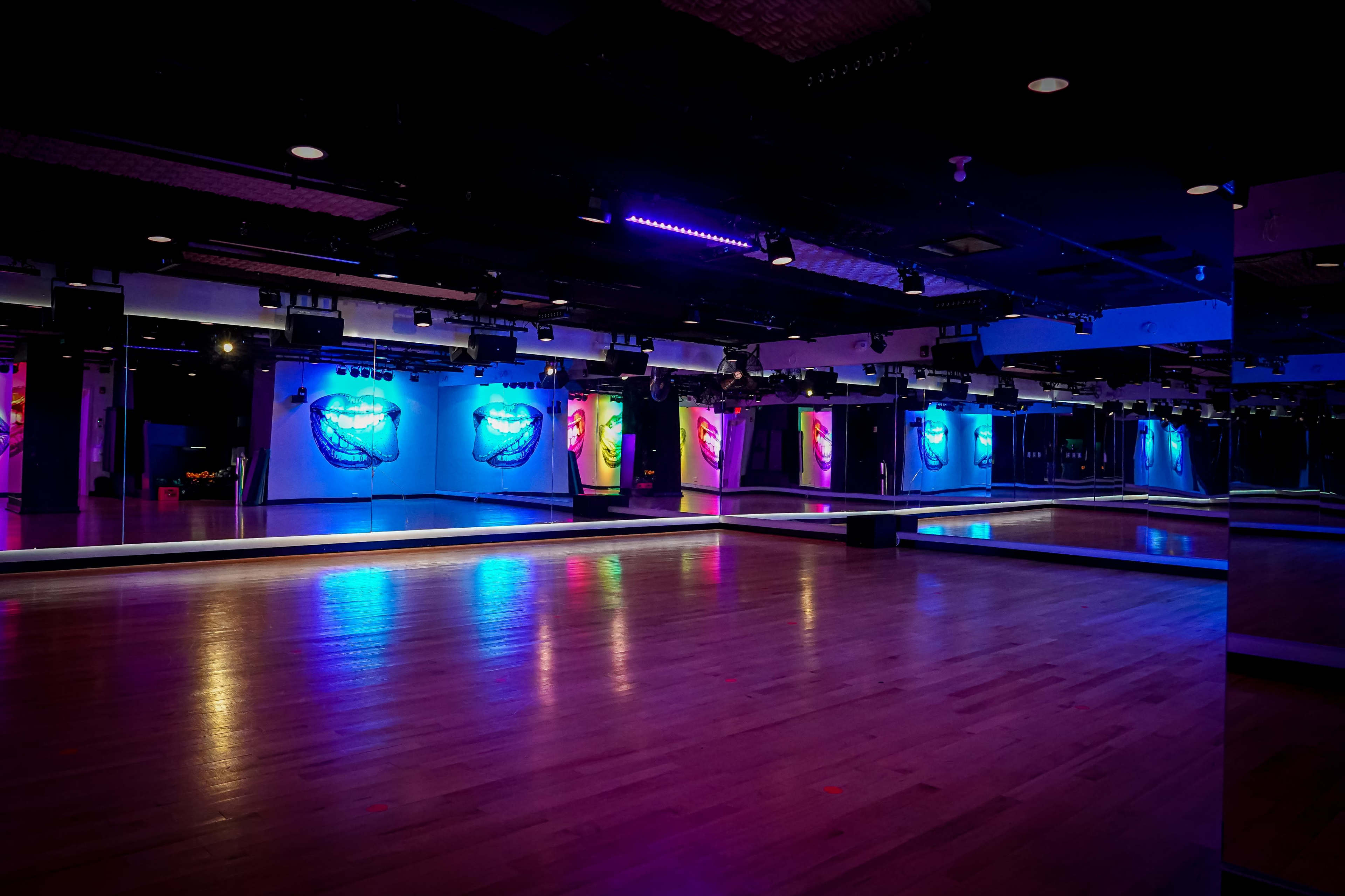 The image shows a dimly lit dance studio with colorful lighting and large mirrors on the walls.