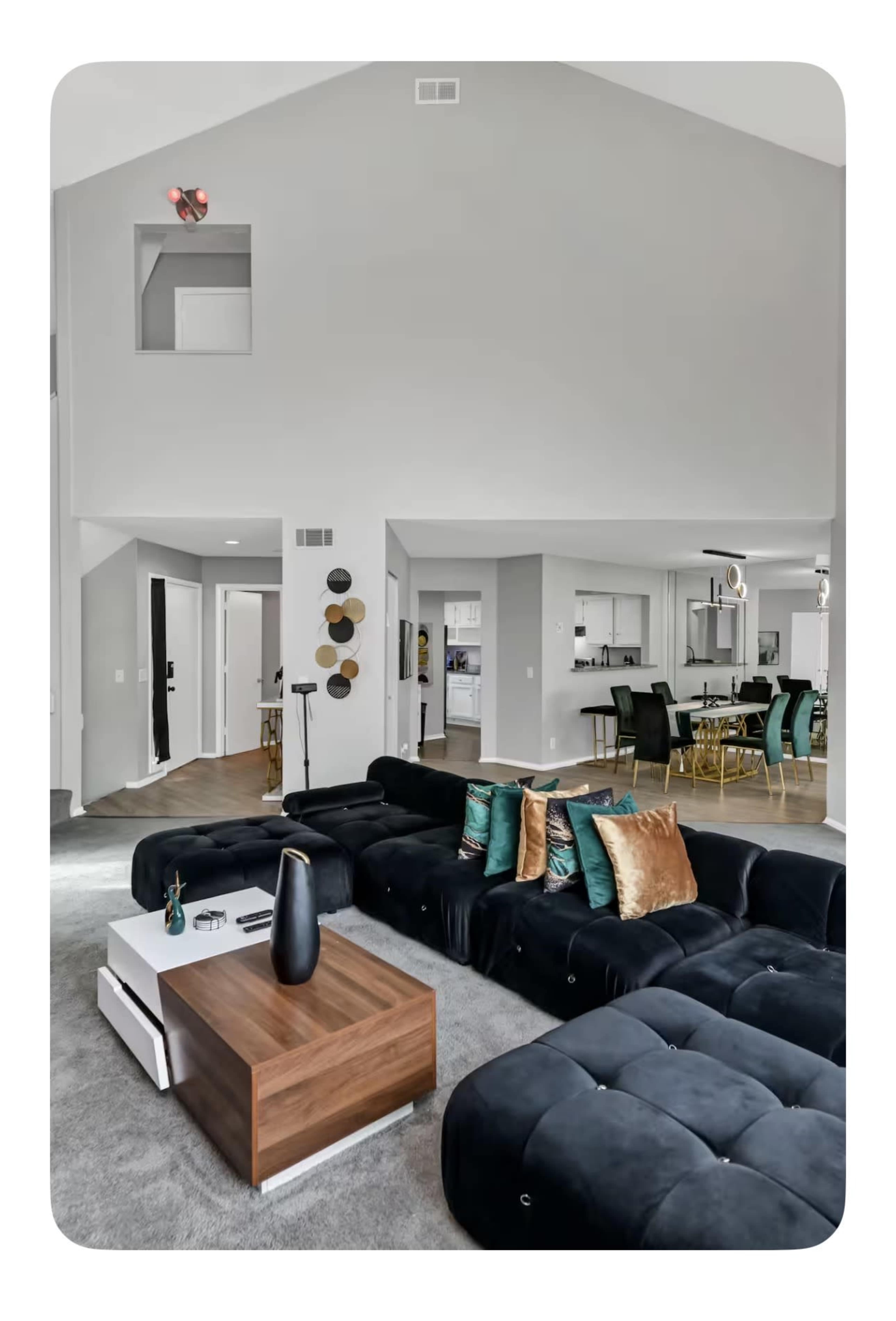 The image shows a modern living room featuring a large black sectional sofa, a wooden coffee table, and contemporary decor with a dining area visible in the background.