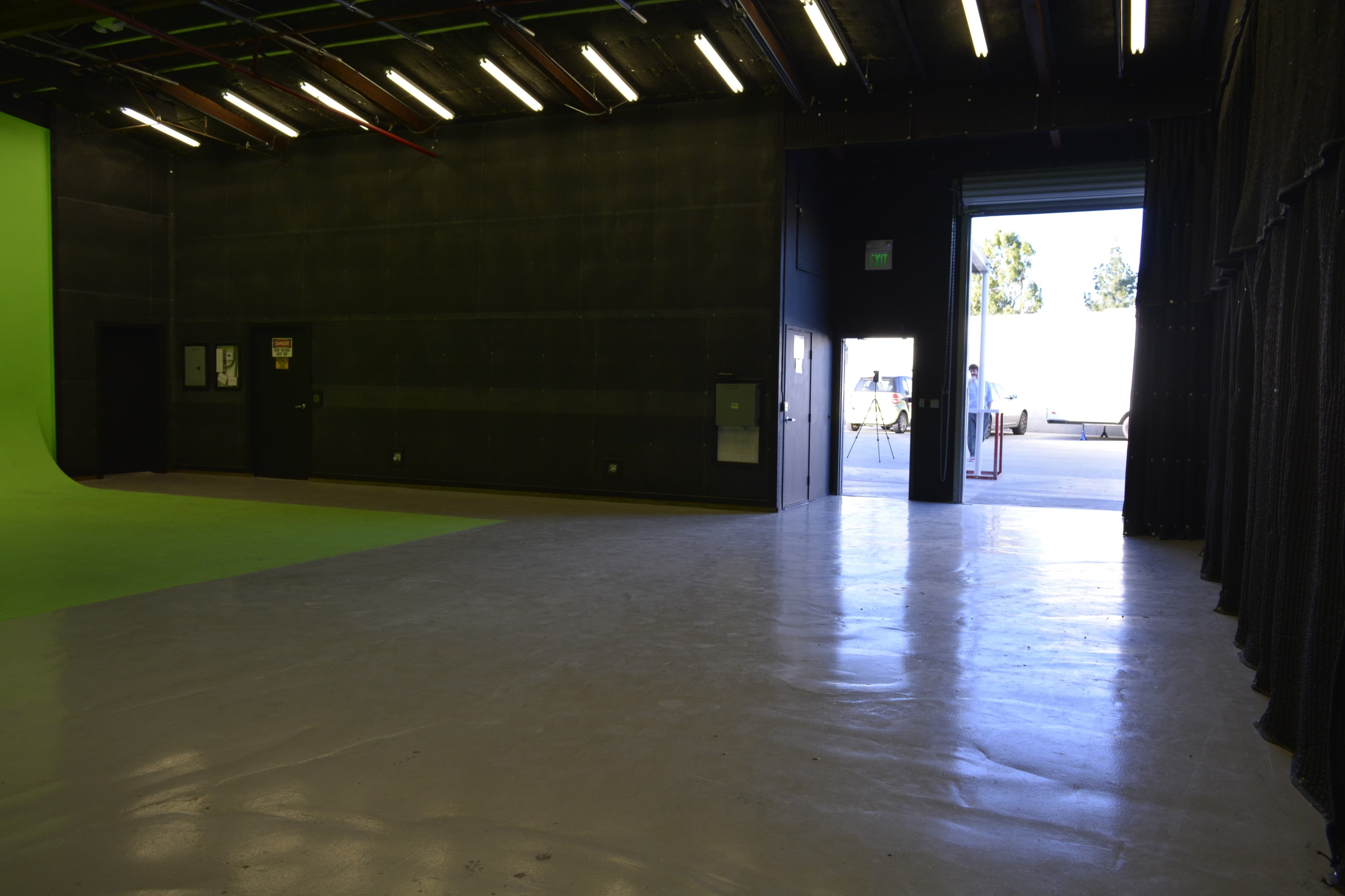 A spacious film studio with a green screen wall on one side and an open door leading outside on the opposite side.