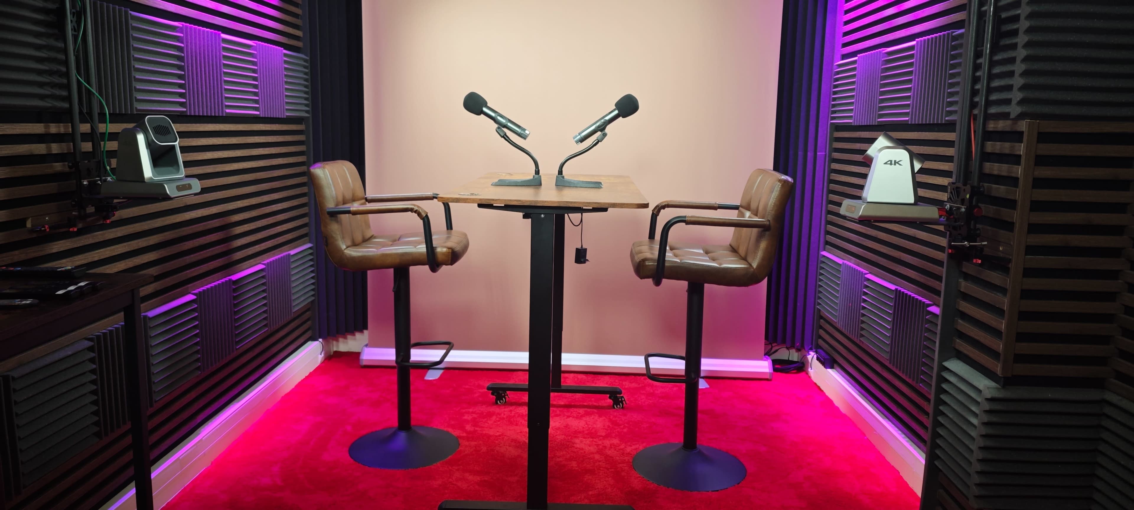 The image shows a podcast recording studio with two high stools, a wooden table, and two microphones positioned in front of a plain backdrop, surrounded by soundproofing panels and ambient lighting.