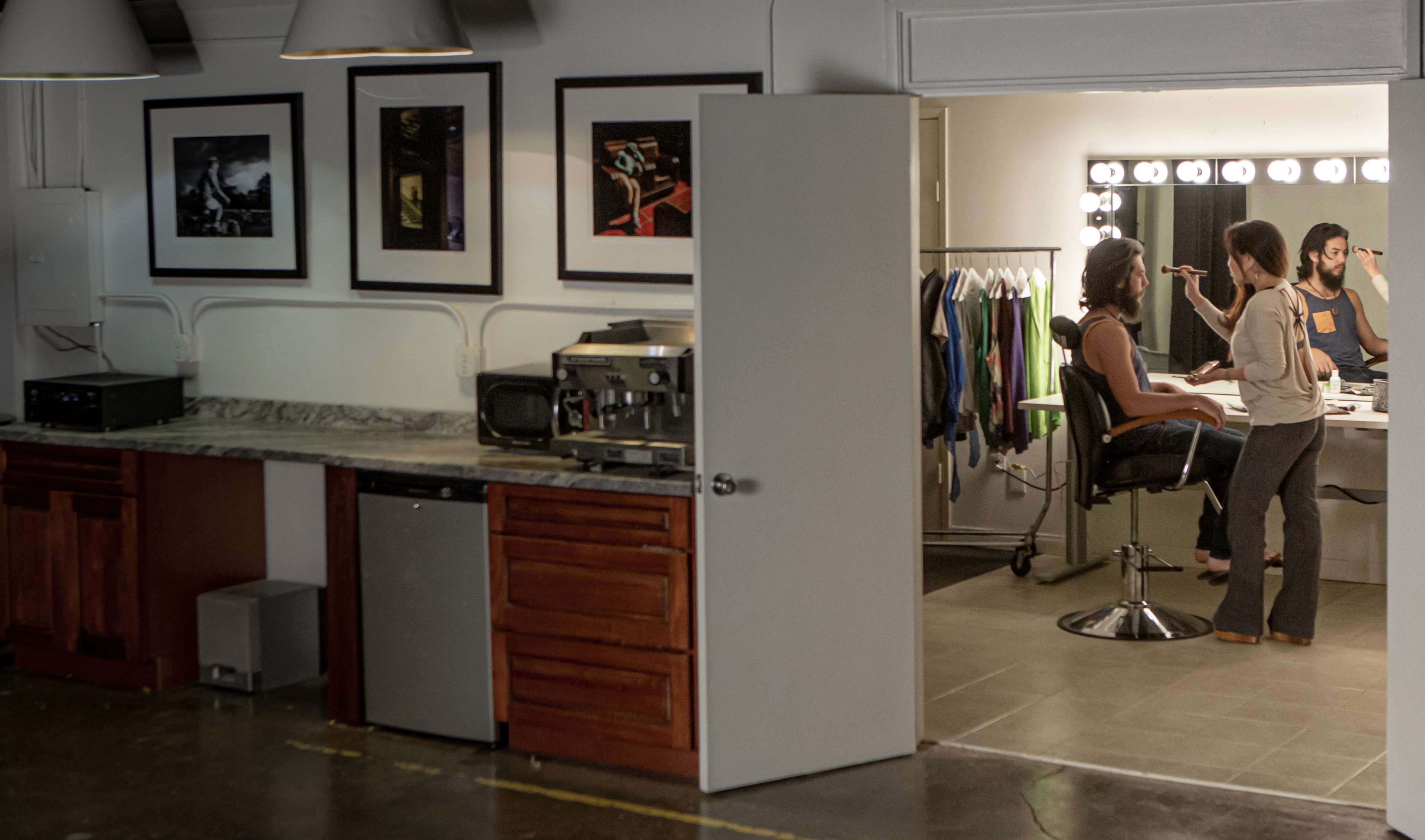 A makeup artist applies makeup to a client at a vanity in a room with a kitchenette and framed pictures on the wall.