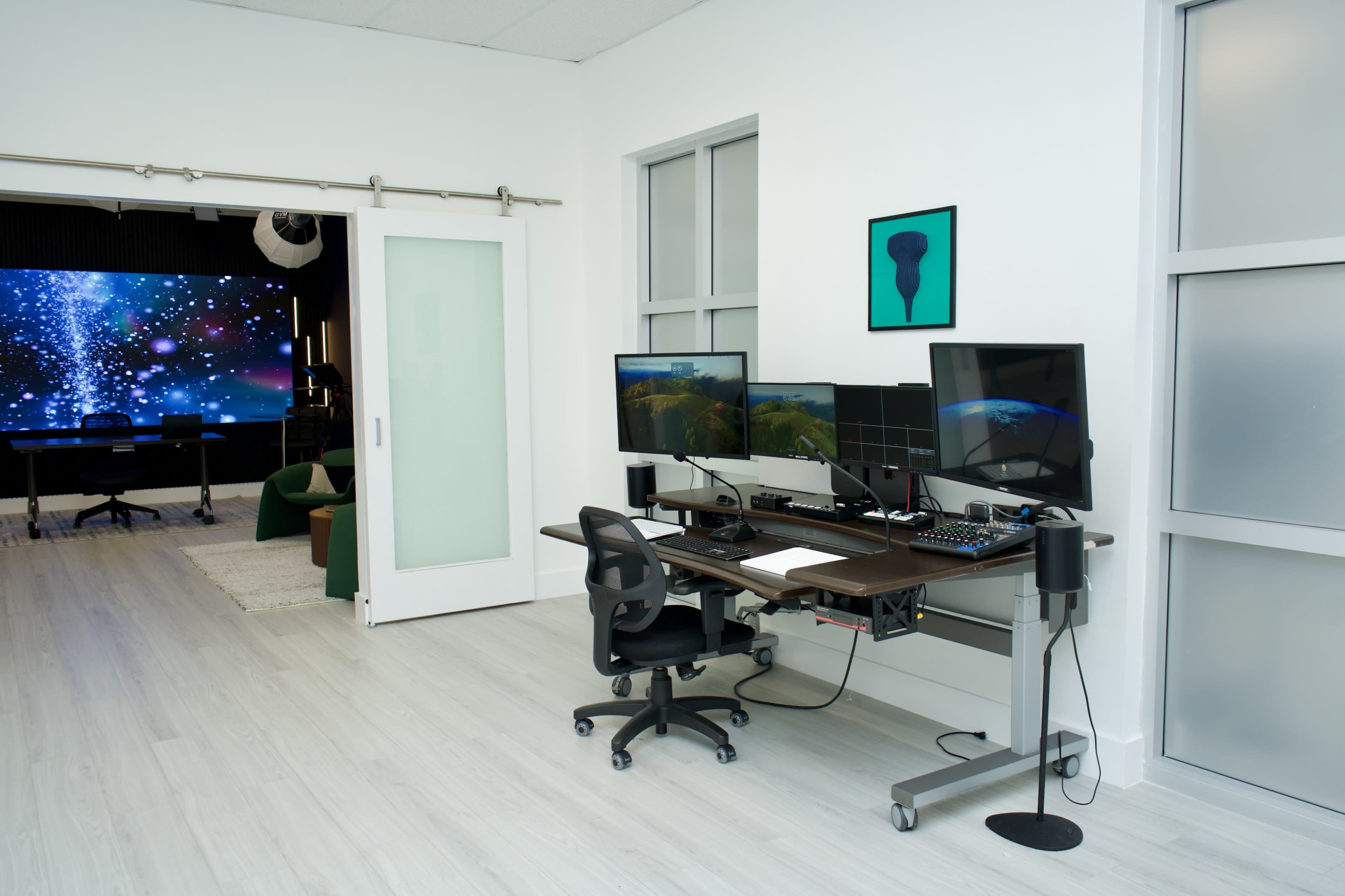 A modern office space features a desk with multiple computer monitors and a clear view of a room with a large screen in the background.