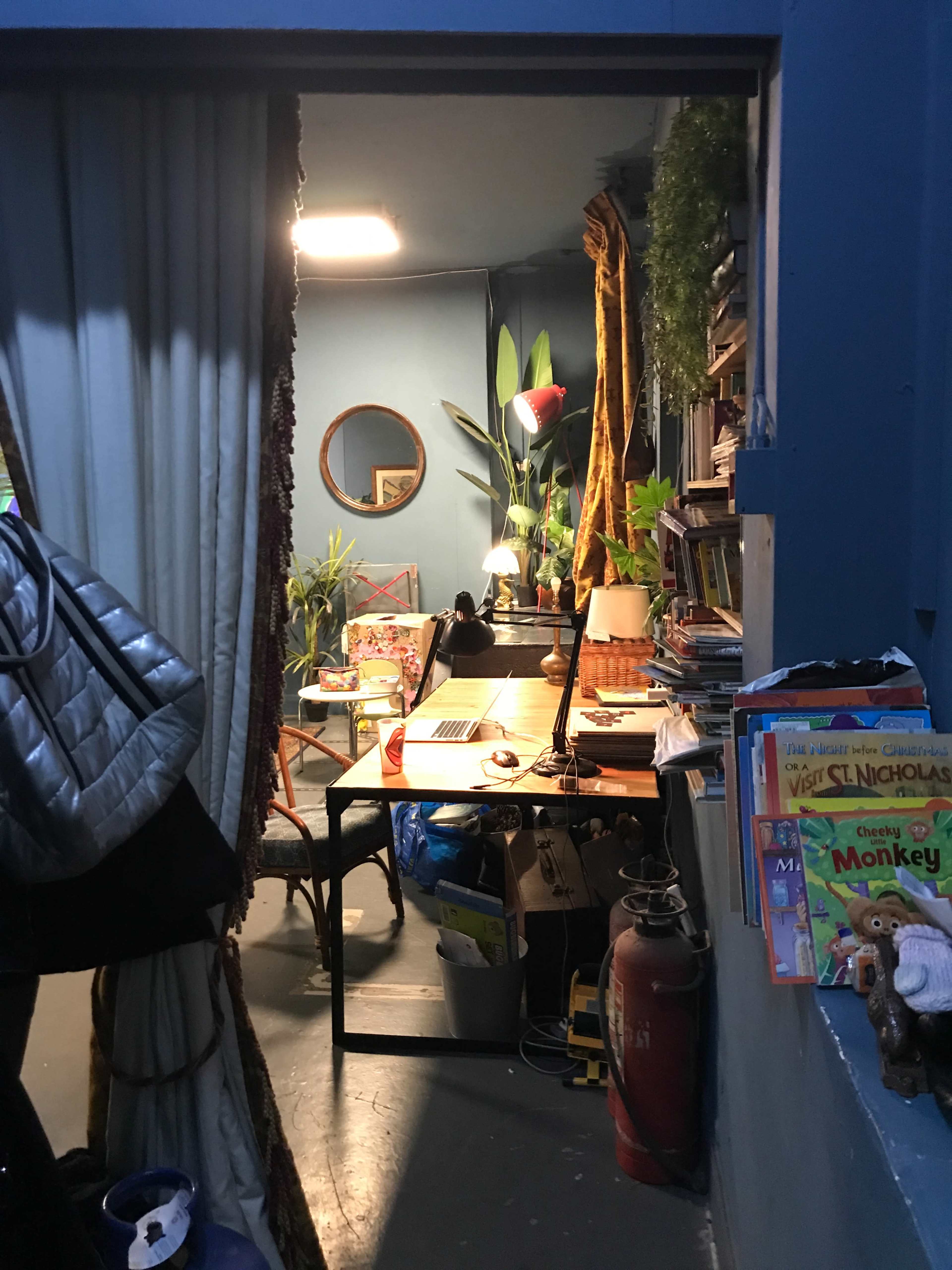 A small room features a desk with a laptop surrounded by plants, bookshelves, and various supplies, with a curtain partially dividing the space.