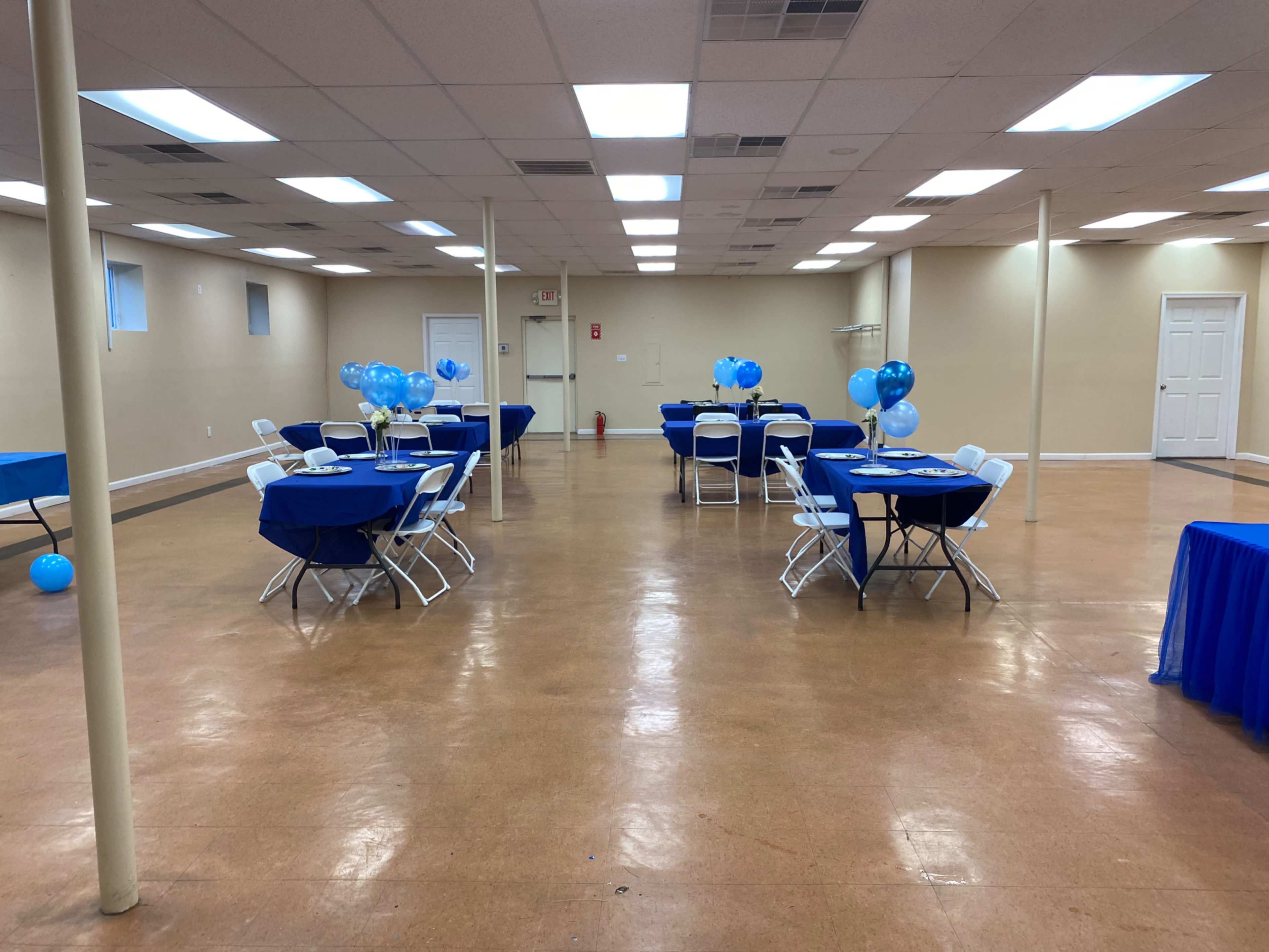 The image shows a spacious event room with blue-tabled seating arrangements and balloons scattered throughout the area.
