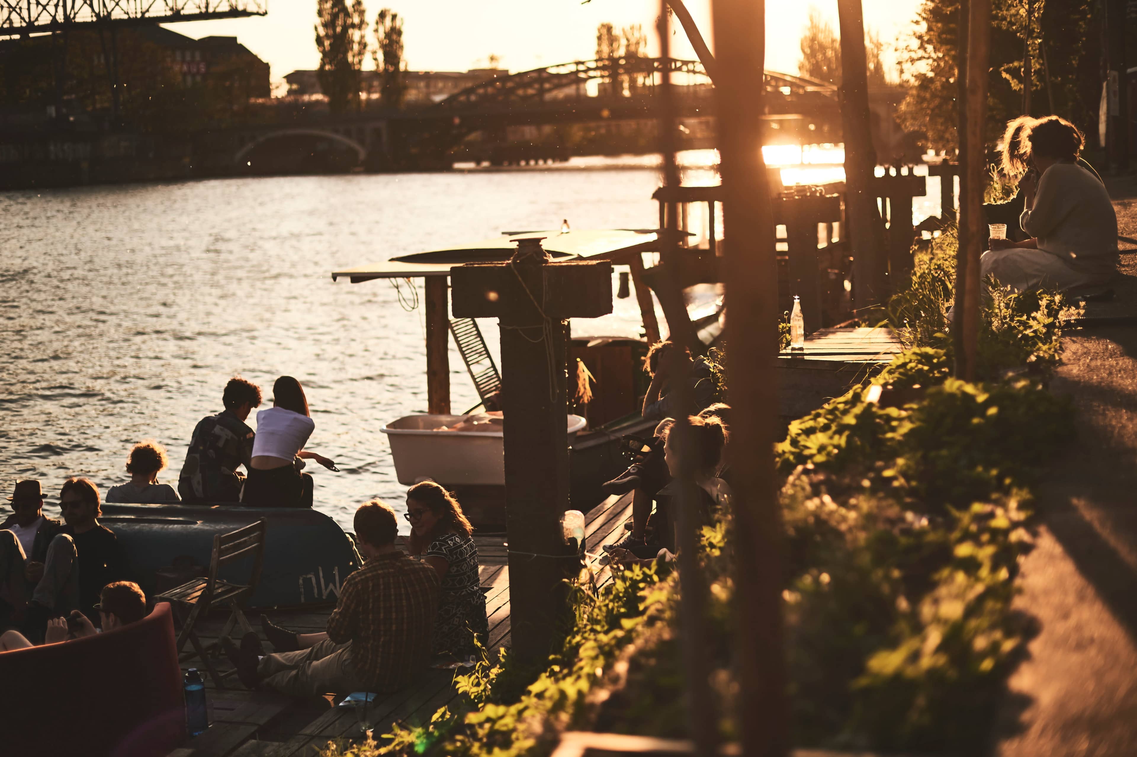 People are gathered along a waterfront, sitting on a dock and socializing as the sun sets.