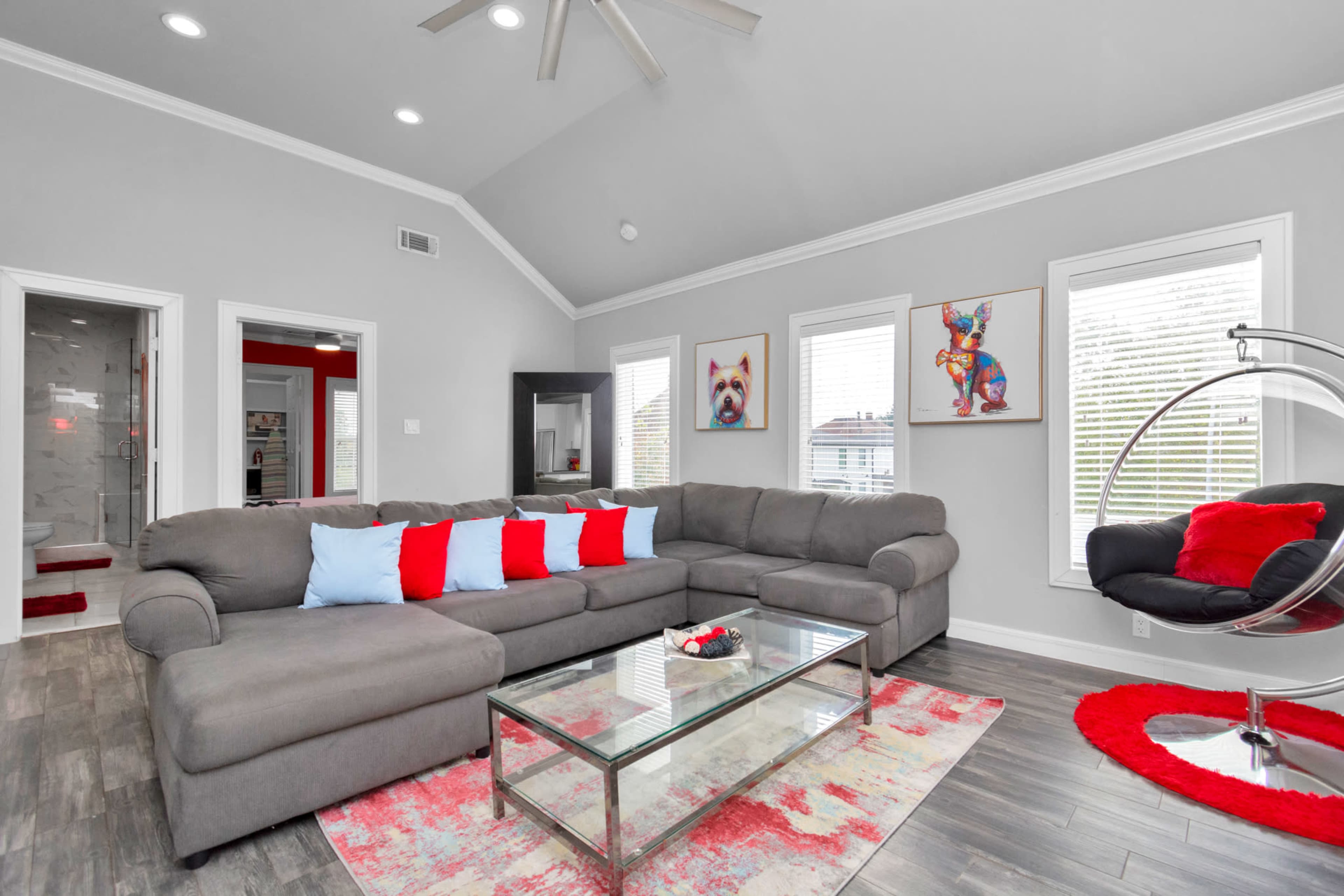 The living room features a spacious gray sectional sofa, a glass coffee table, and vibrant artwork on the walls, with large windows allowing natural light to fill the space.