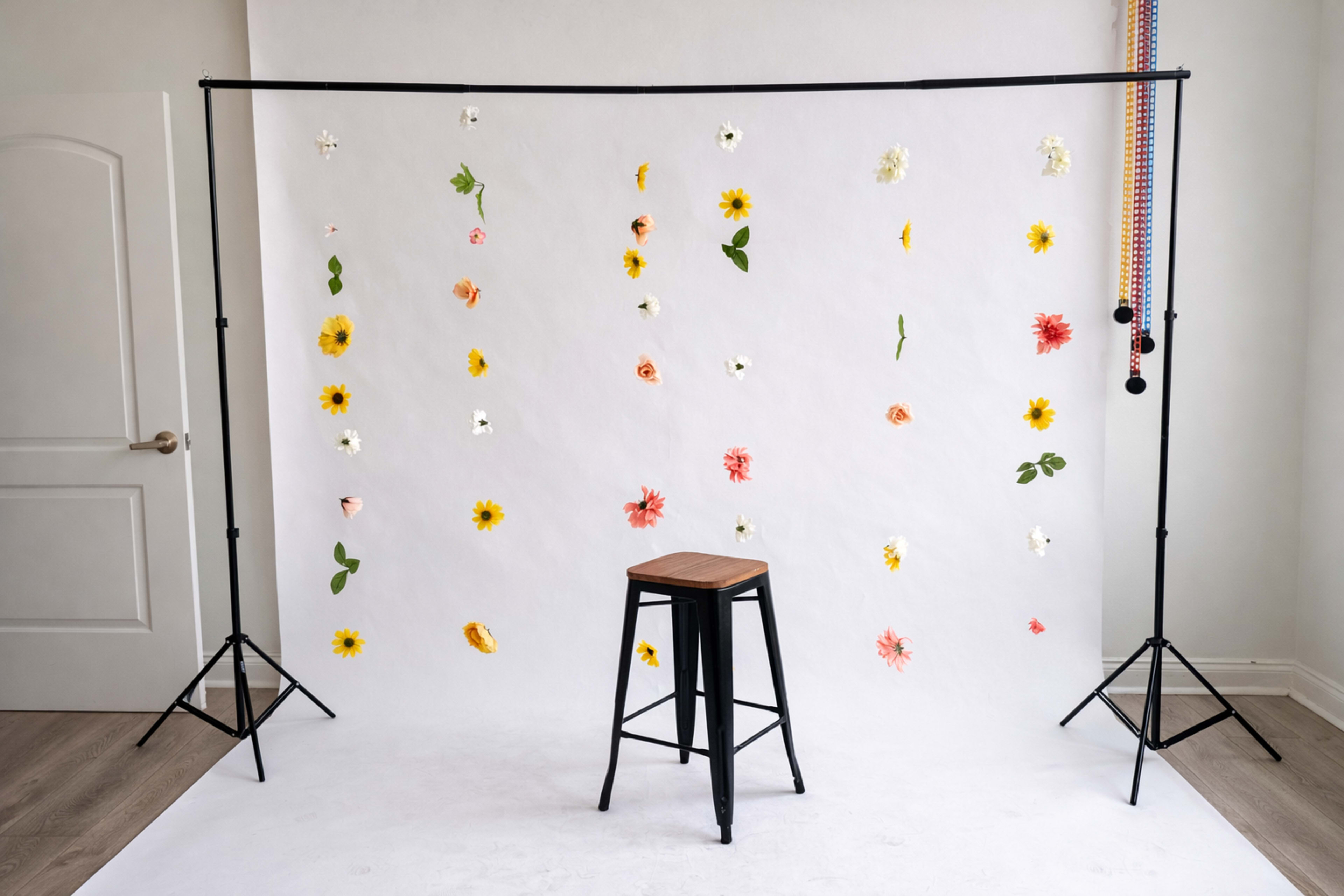 The image shows a photography backdrop with a white paper surface decorated with scattered flowers, alongside a black stool positioned in front of it.