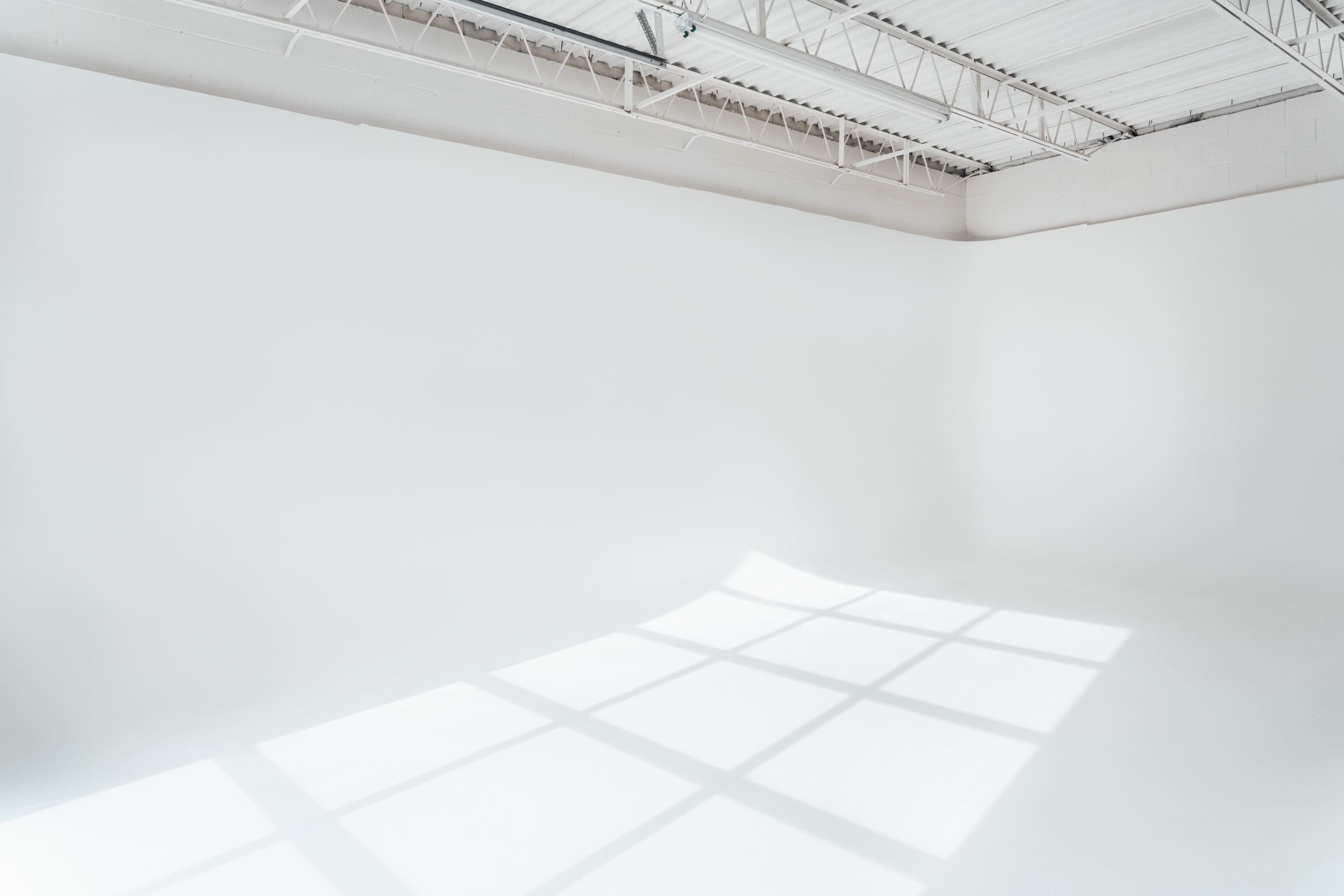 A large, empty studio space with white walls and a bright, illuminated floor creating shadows from window grids above.