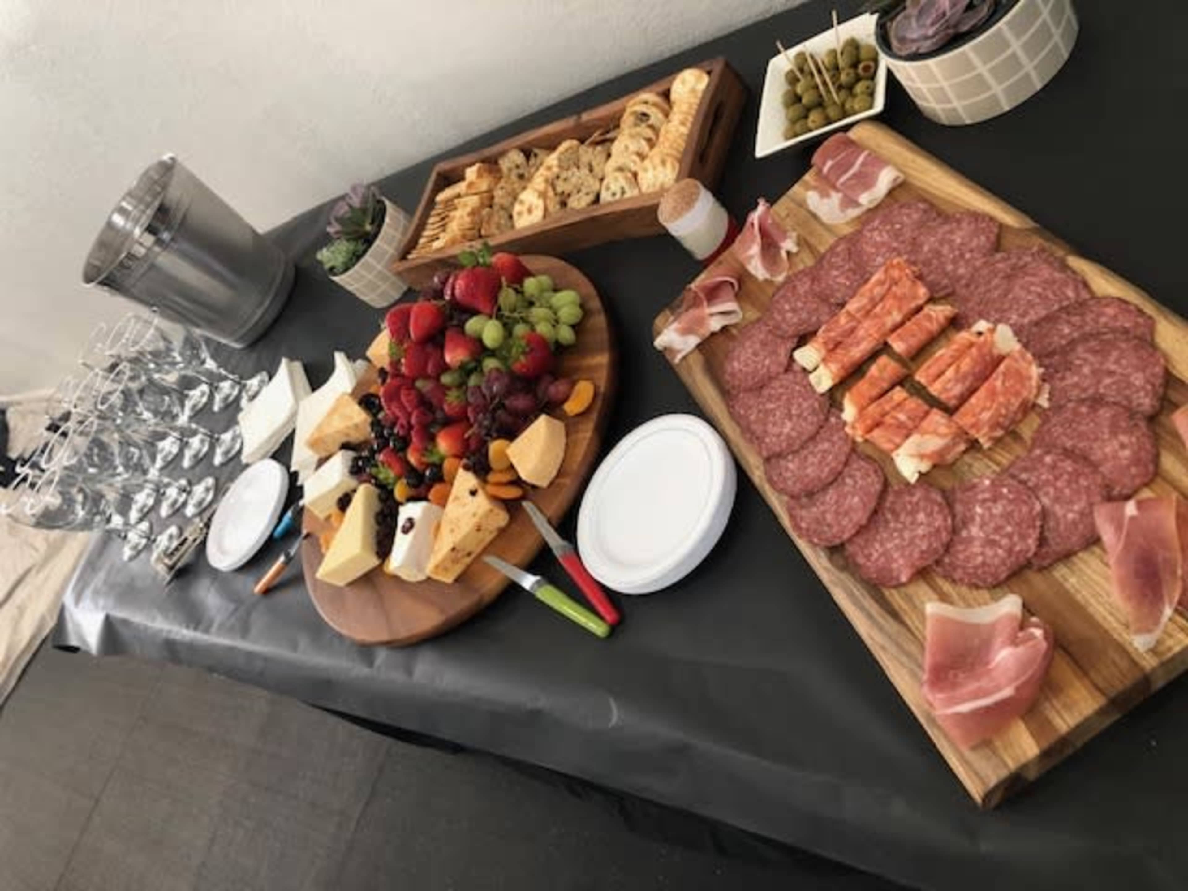 A table is set with a variety of cheeses, cured meats, fruits, nuts, crackers, and glasses, alongside bowls of olives and garnishes.