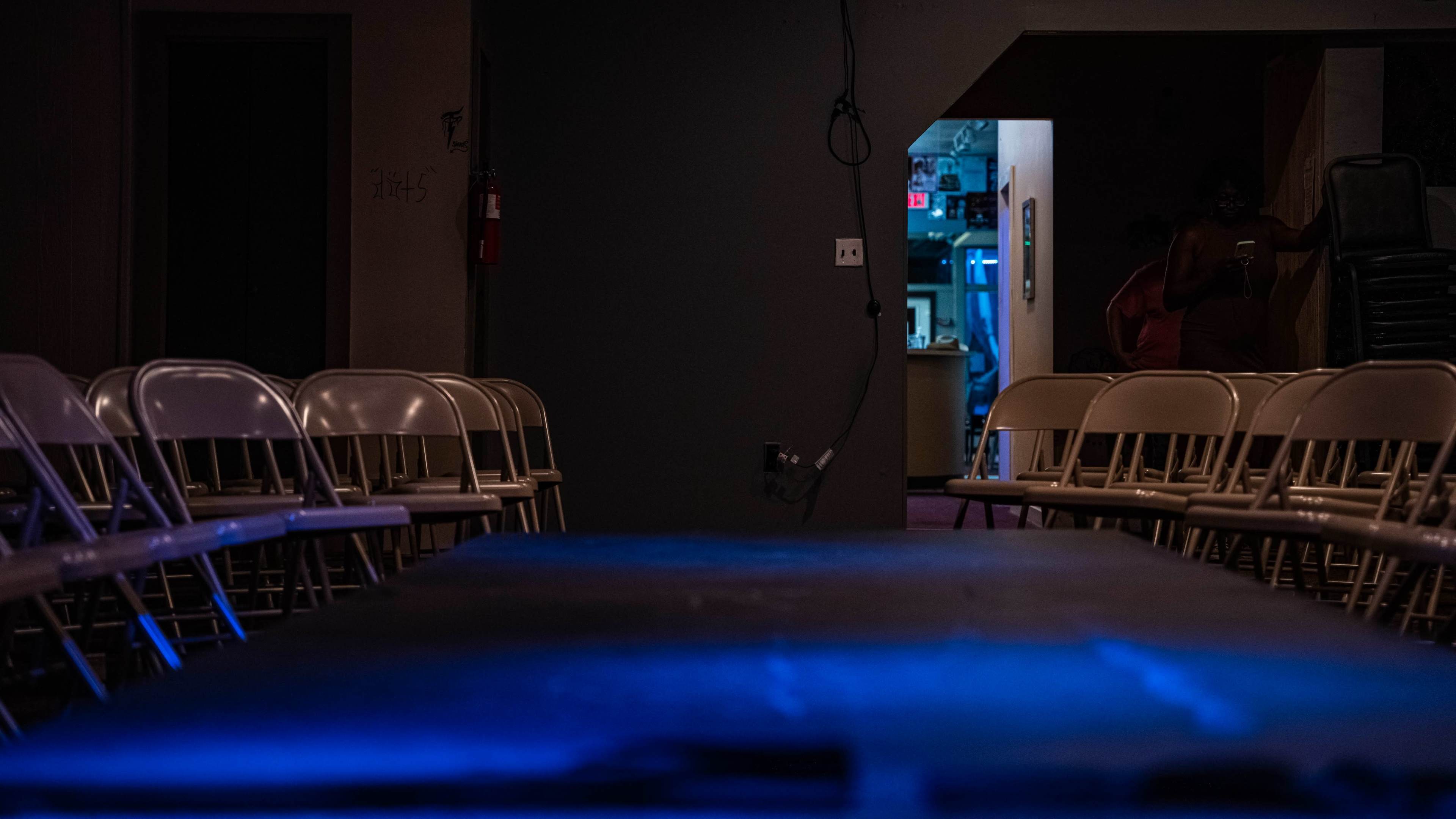 A dimly lit room with rows of white chairs facing a table, and a doorway leading to a brighter area in the background.