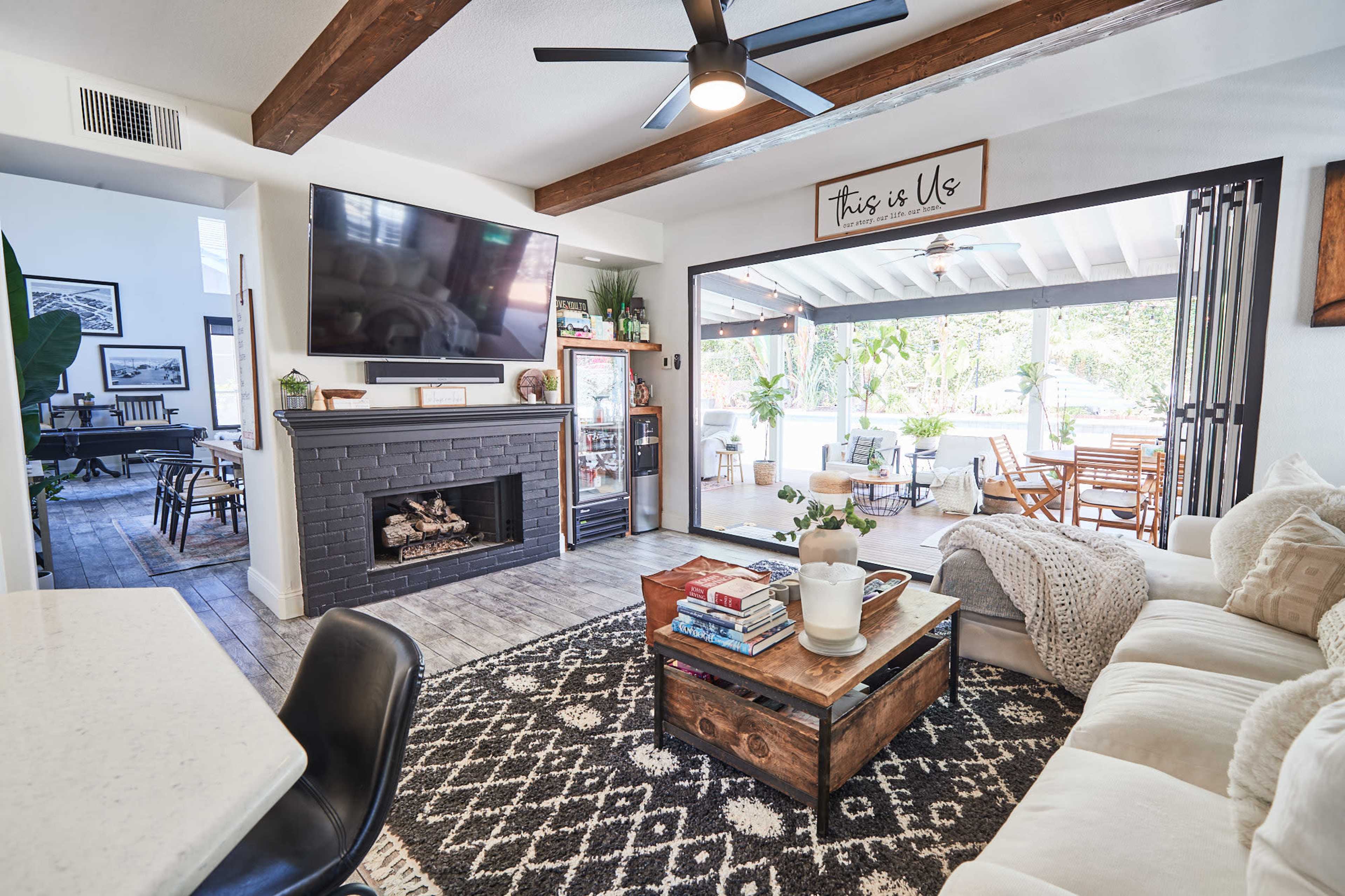A living room features a modern fireplace, a large television, and a coffee table, with a view of an outdoor seating area through open glass doors.