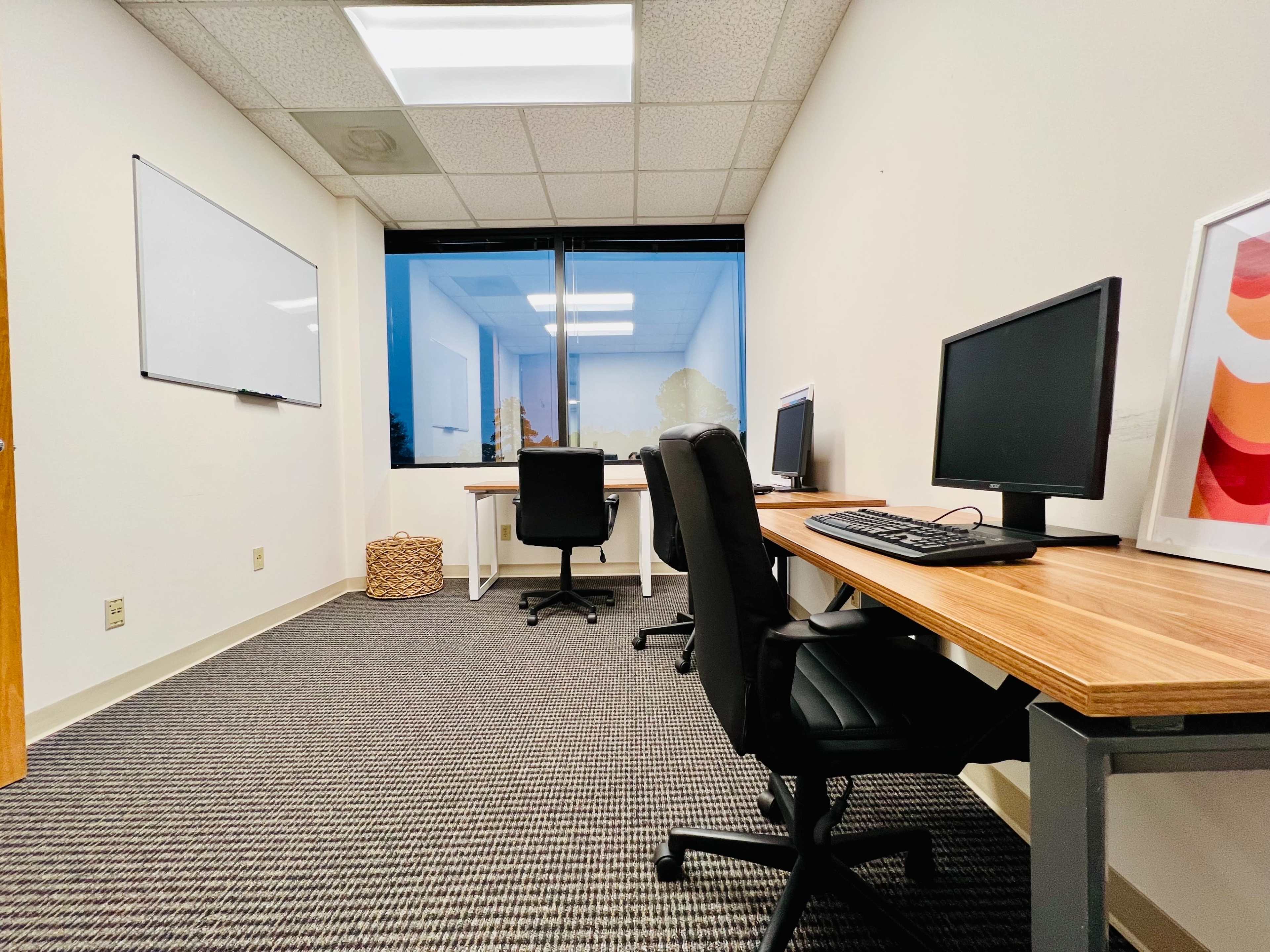 A sparsely furnished office space features three chairs and two computer desks, with a whiteboard on the wall and a large window displaying an outdoor view.