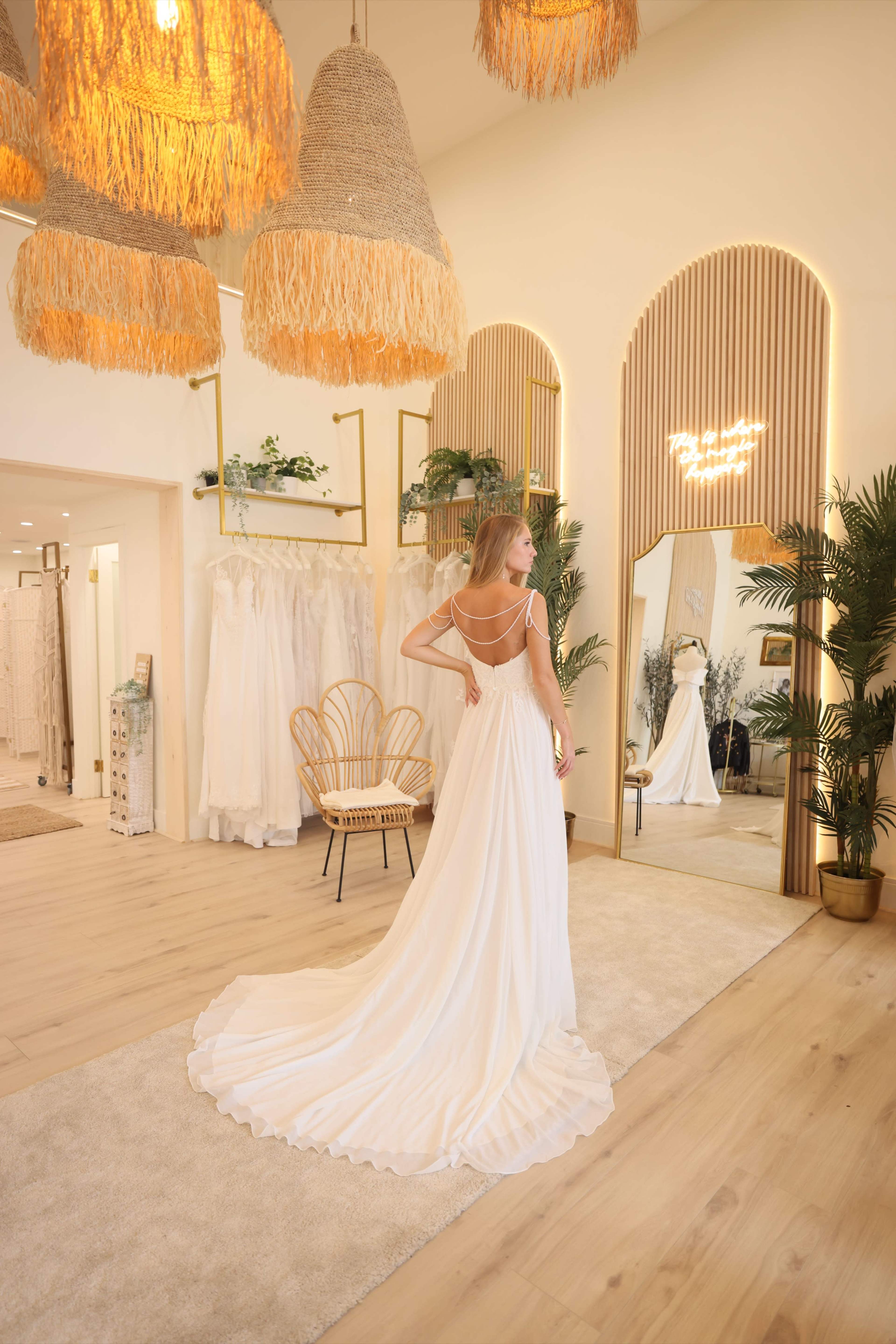 A woman wearing a flowing white gown stands in a boutique adorned with woven light fixtures and a mirrored wall.
