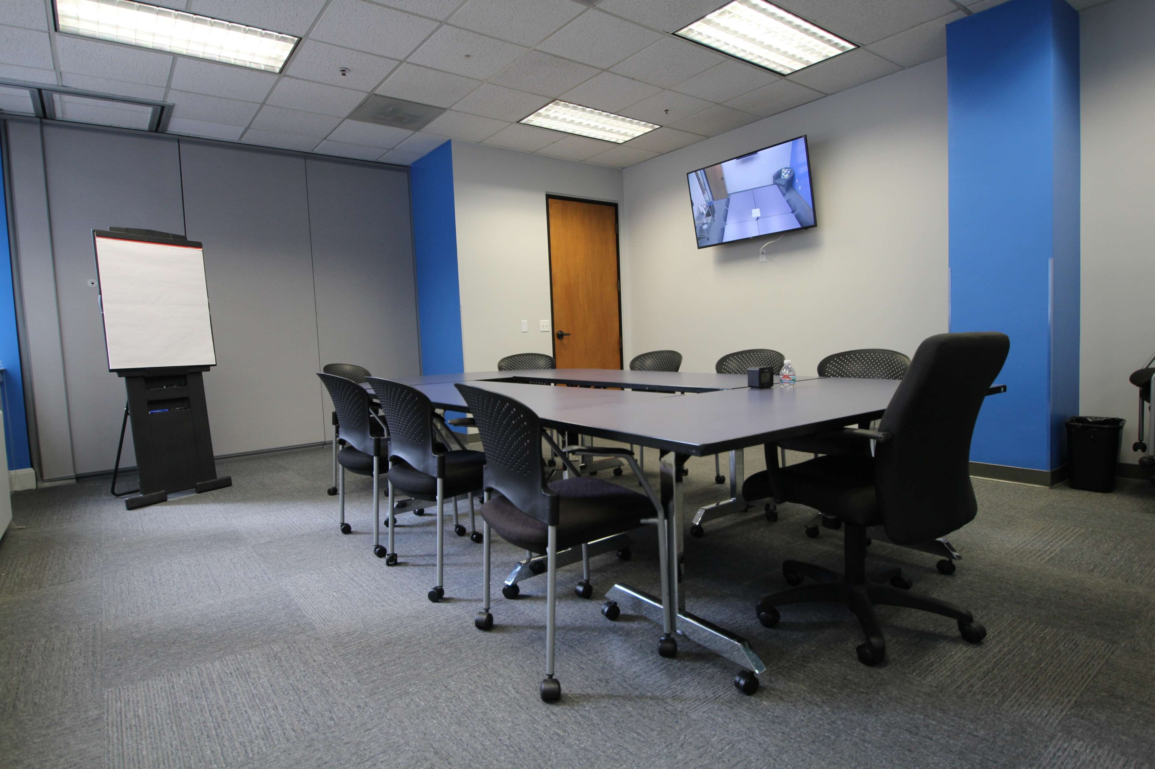 A modern conference room features a large table surrounded by black ergonomic chairs, a whiteboard, and a wall-mounted television.