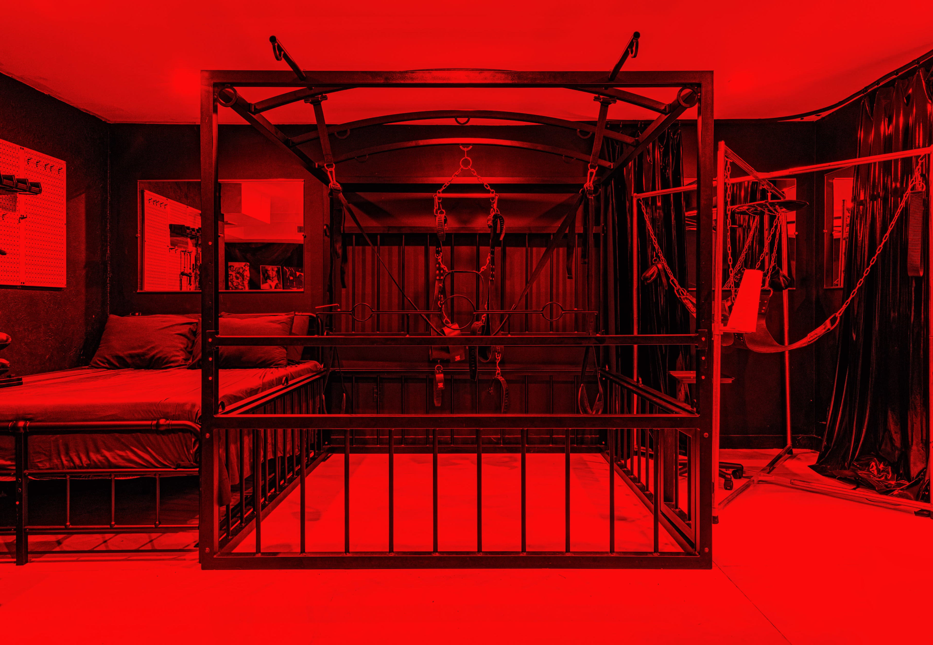 A red-lit room featuring a metal-frame bed with a surrounding bondage frame equipped with restraints.