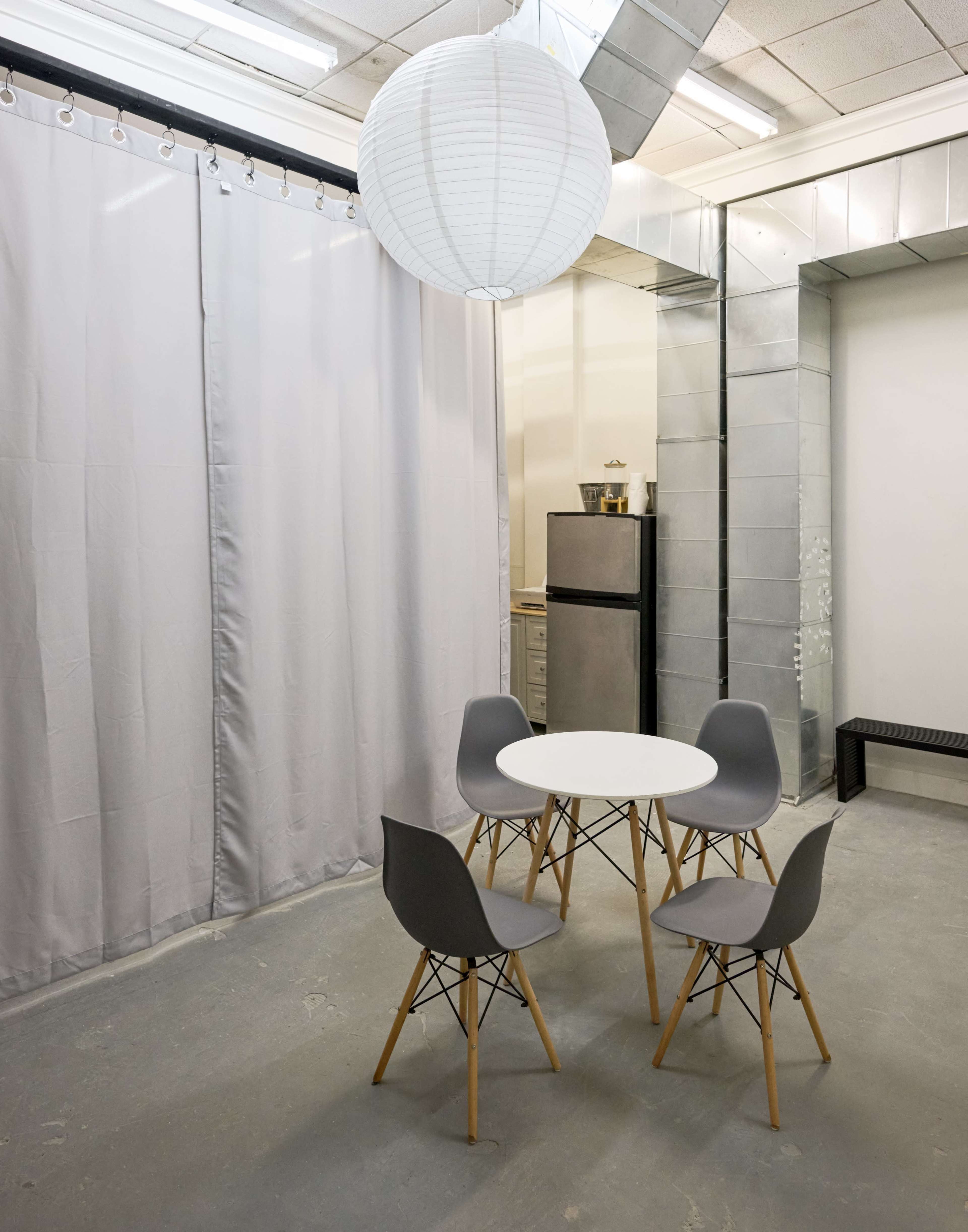 The image shows a minimalist room with a round white table and four gray chairs, a refrigerator in the background, and a paper lantern hanging from the ceiling.