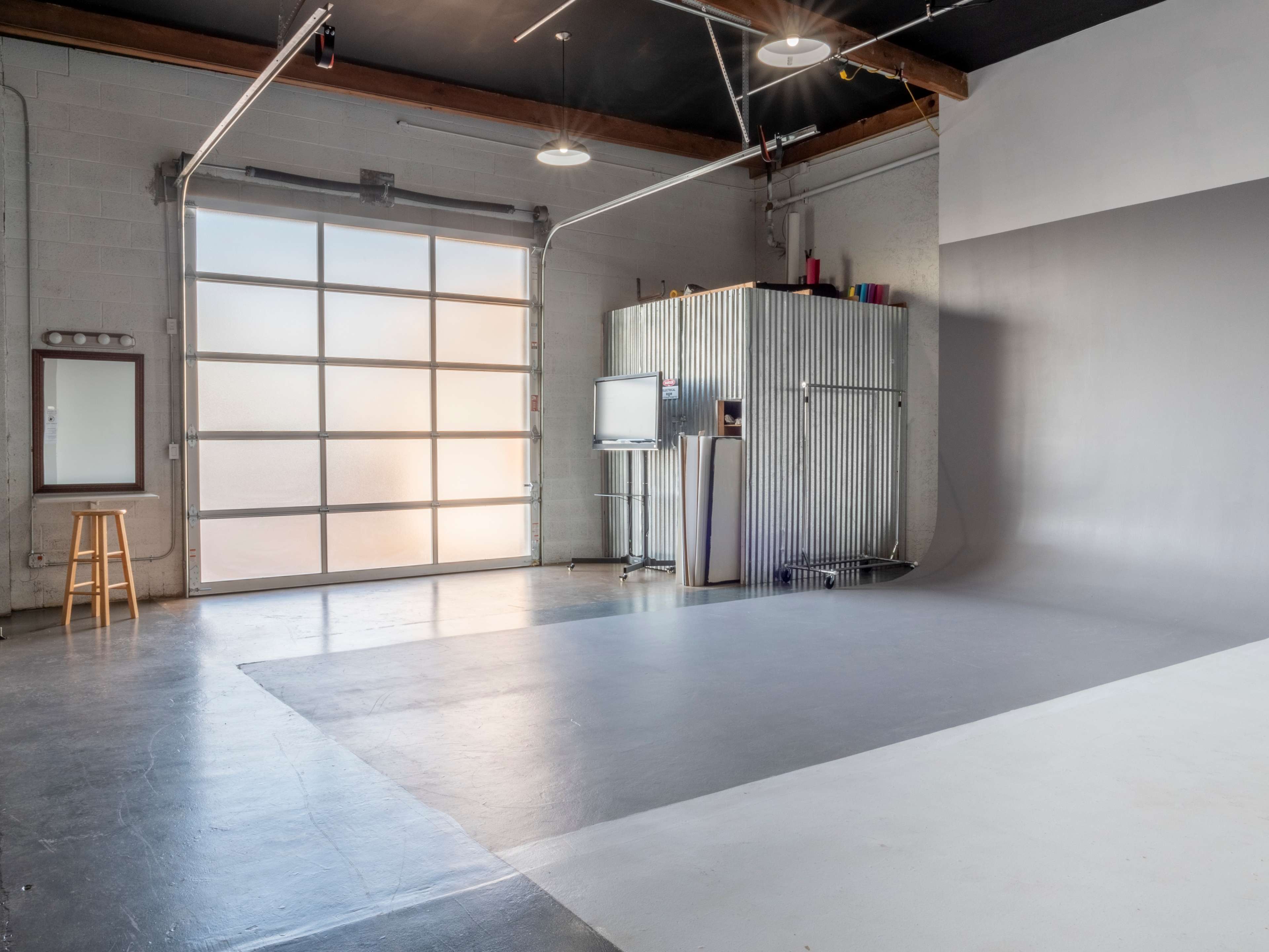 The image shows a spacious studio with a large garage door, a metal storage unit, a flat screen TV, and a bare, light gray backdrop.