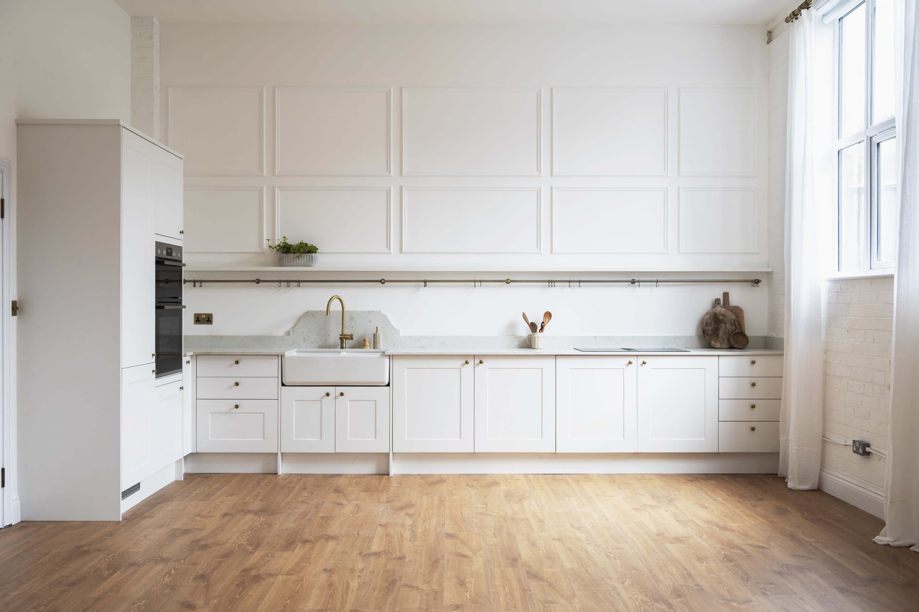 The image shows a modern kitchen with white cabinetry, a farmhouse sink, and a wooden floor, featuring a simple countertop.