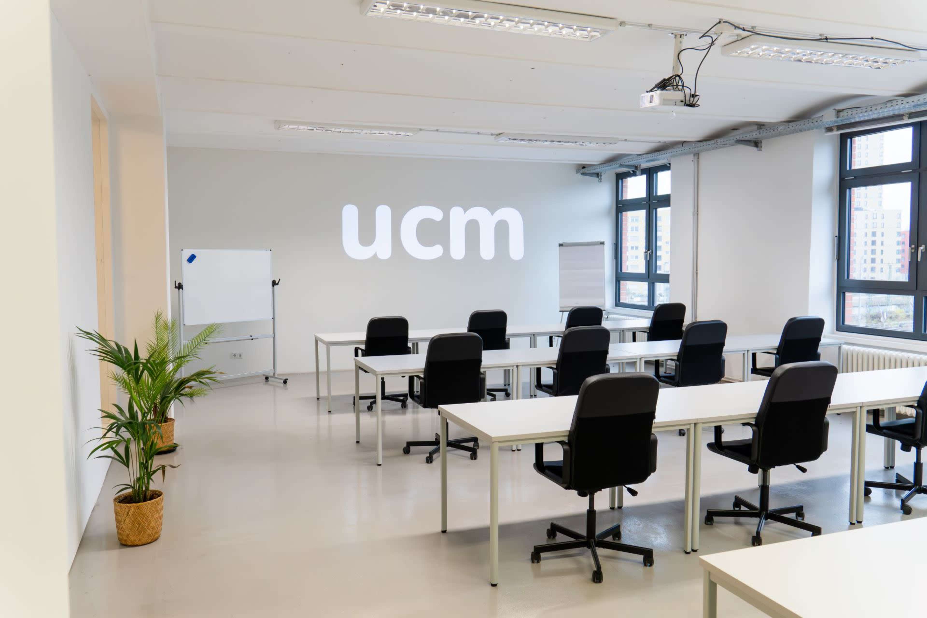 A modern classroom is arranged with several white tables and black chairs, featuring a projector and a large "ucm" logo on the wall.