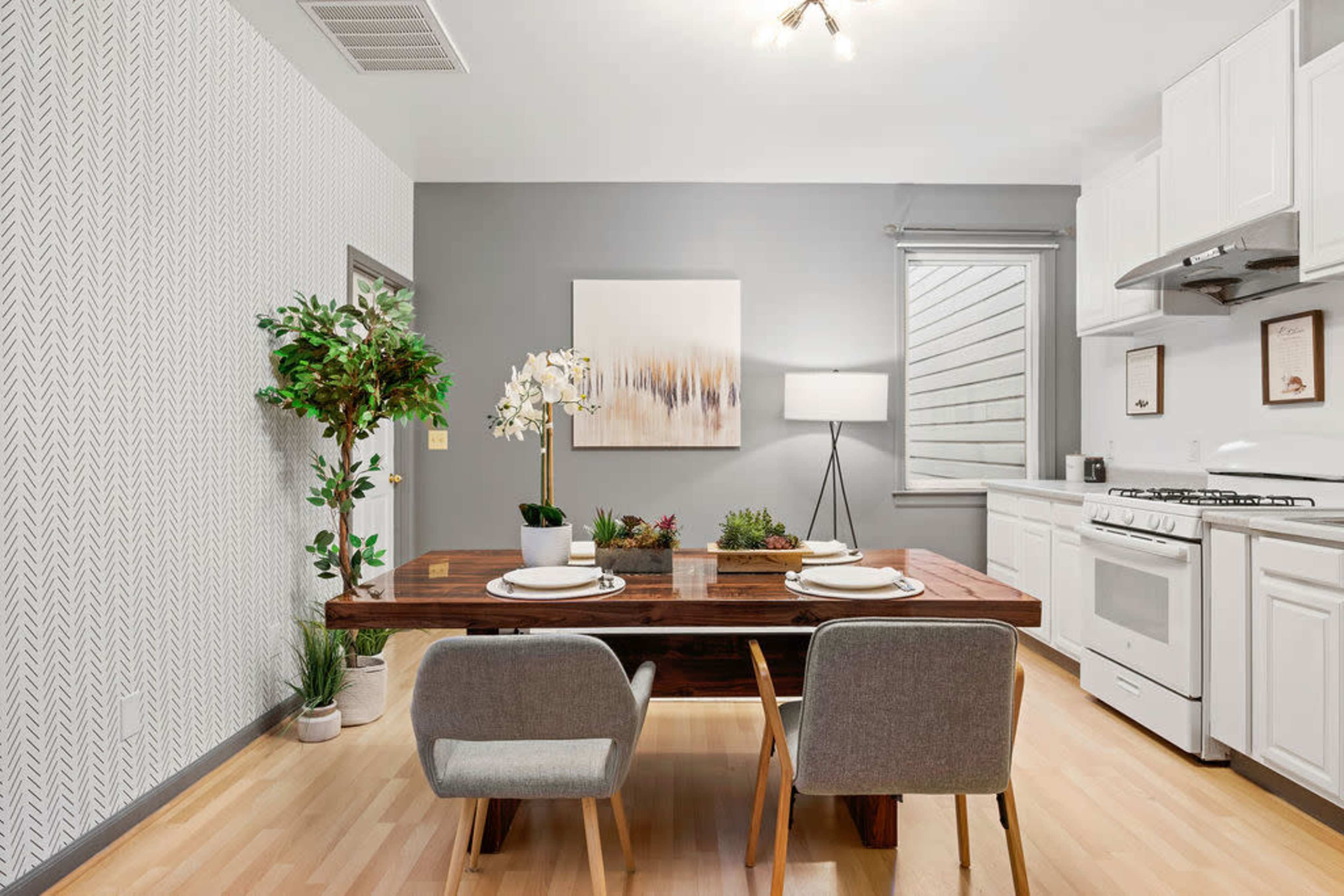A modern kitchen and dining area features a wooden table with two gray chairs, potted plants, and a light fixture overhead, complemented by a light gray wall with a piece of wall art.