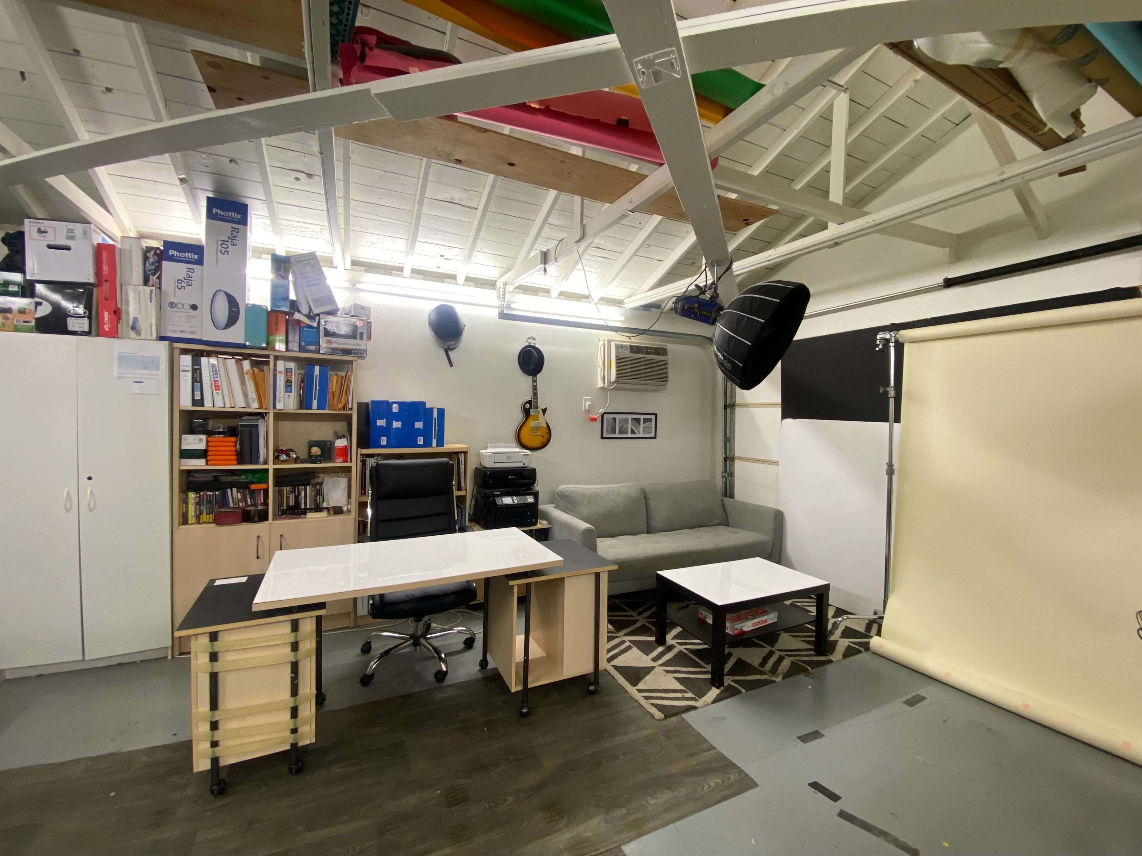 A workspace with a desk, office chair, a gray couch, a coffee table, and various storage items on shelves in a well-lit room.