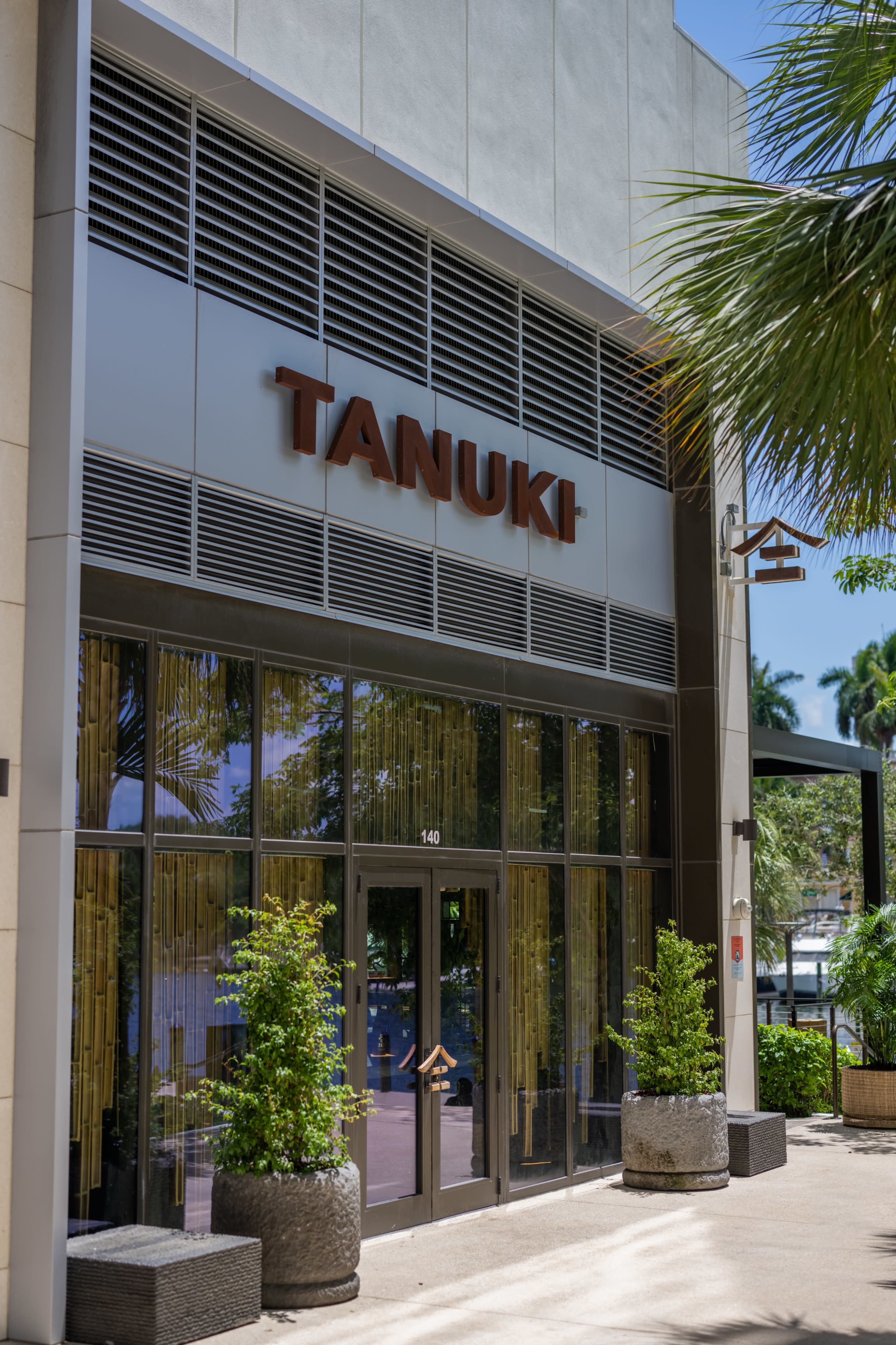 The entrance of a restaurant named "TANUKI," featuring large glass doors and decorative plants in stone planters.