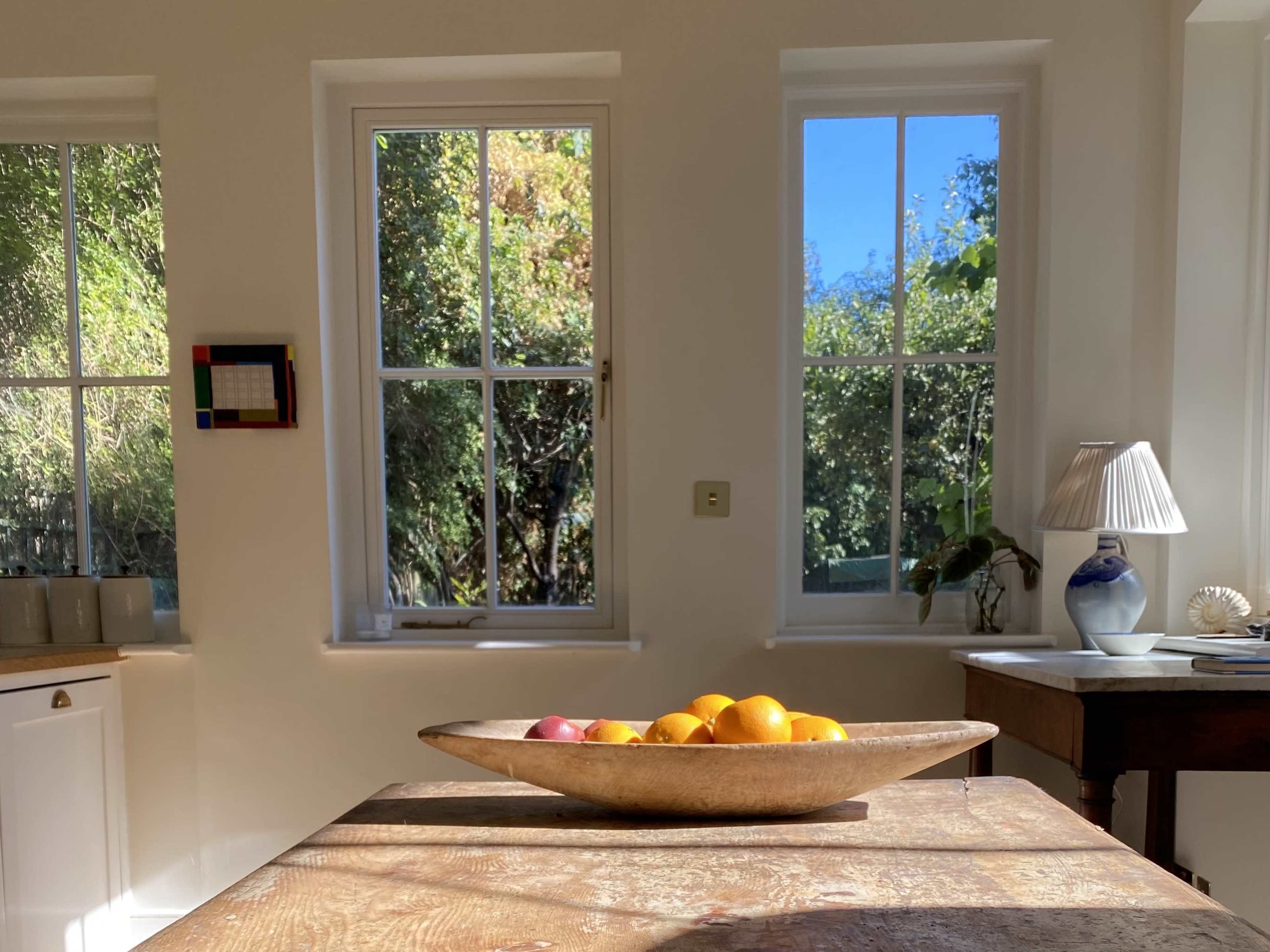 A wooden table holds a bowl of fruit, with sunlight streaming through two windows beside it, illuminating the room.