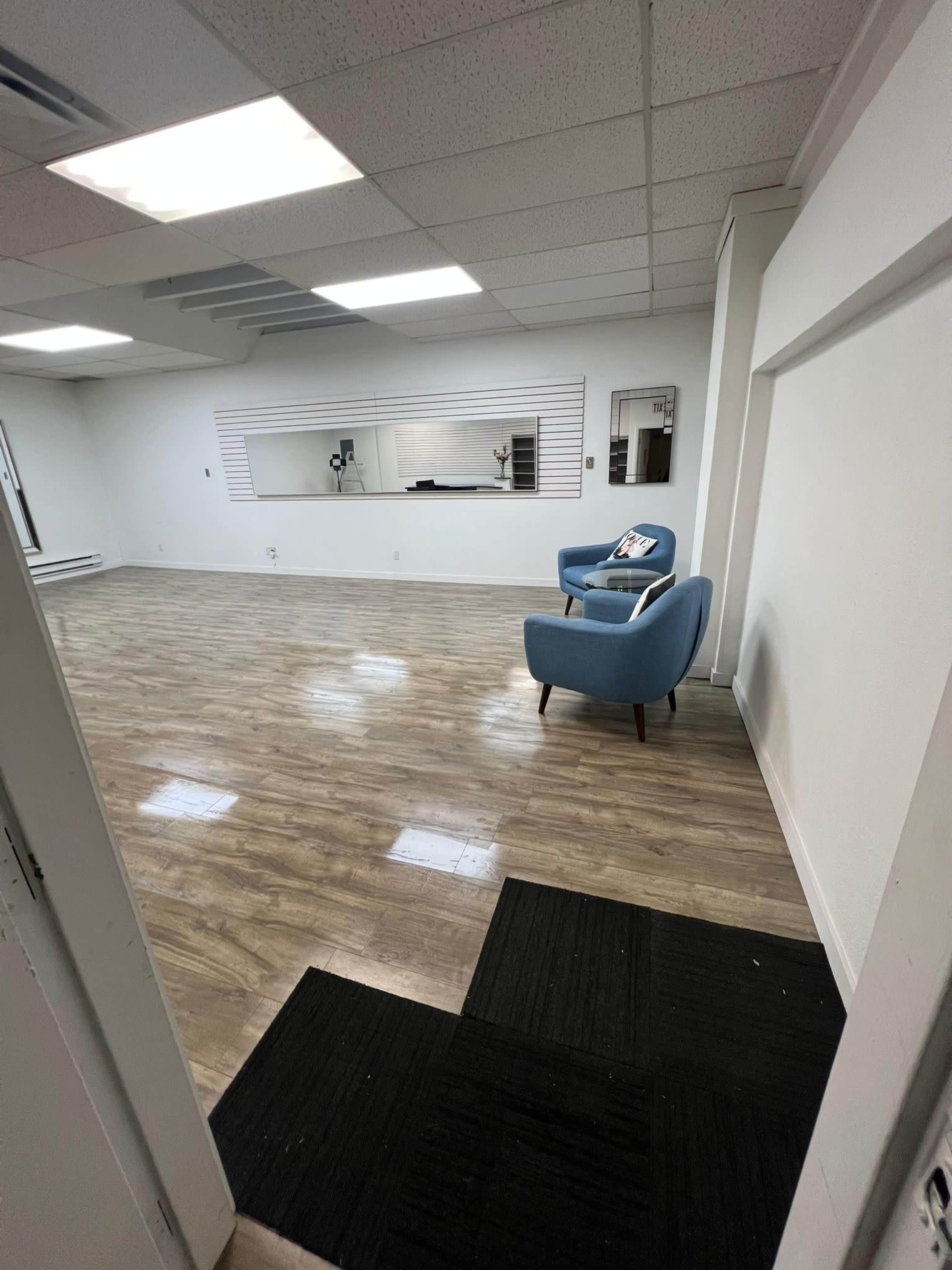 The image shows an empty room with wooden floor tiles, a blue armchair, and a wall-mounted mirror.