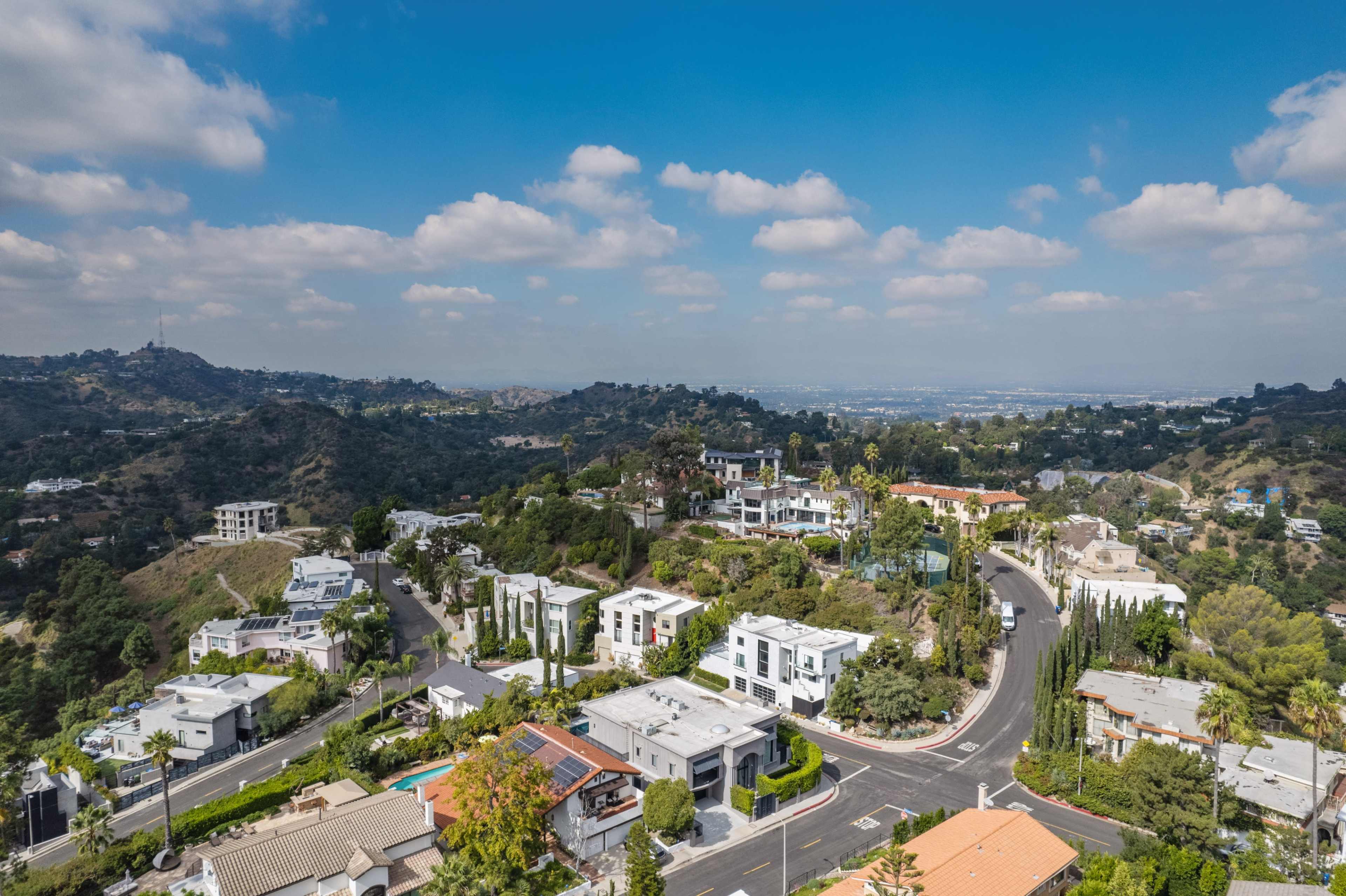 The image shows a panoramic view of a hilly residential area with modern homes and winding roads under a partly cloudy sky.
