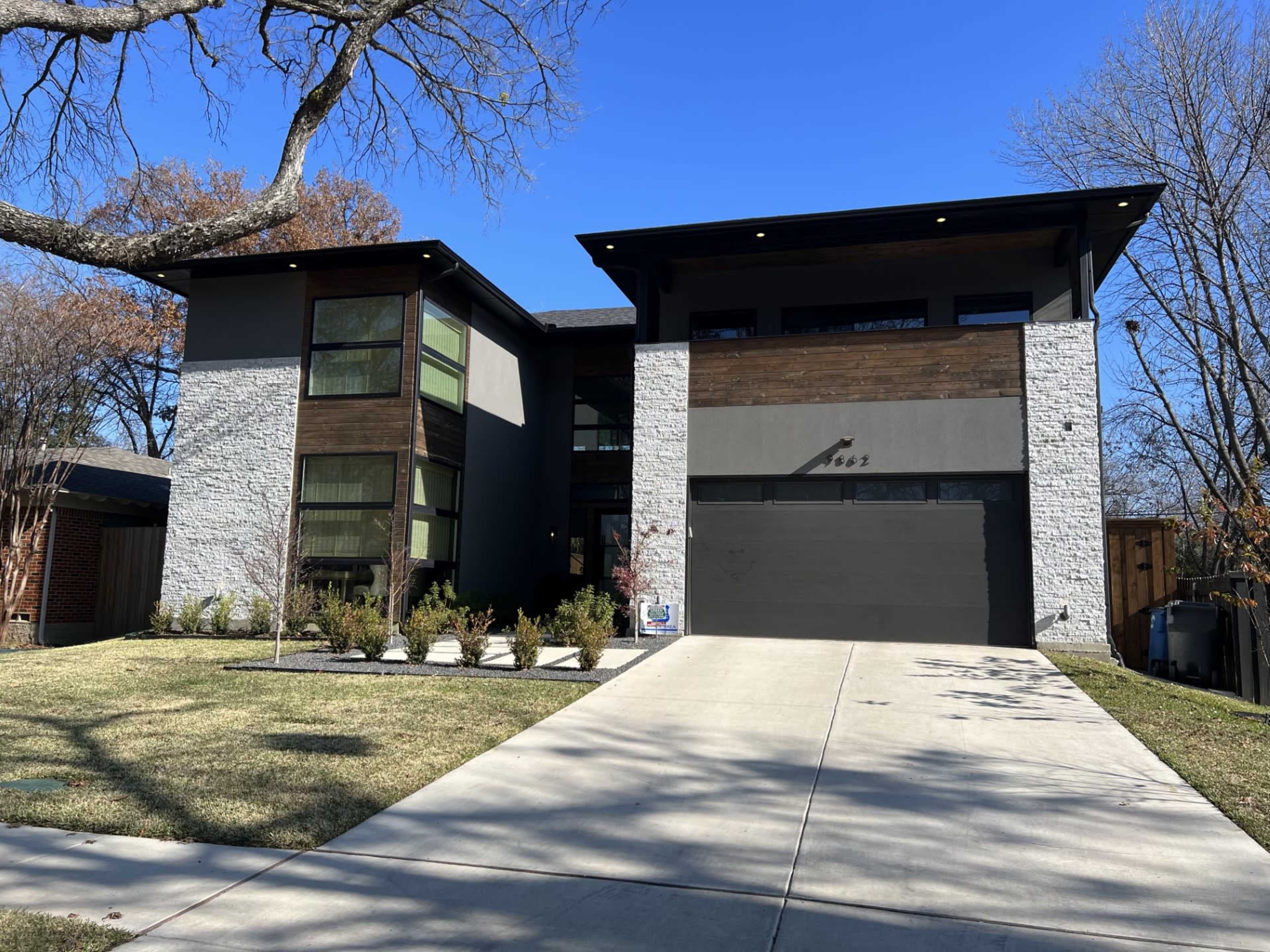 A modern two-story house with a mix of stone and wooden exterior, a large front lawn, and a concrete driveway leading to a garage.