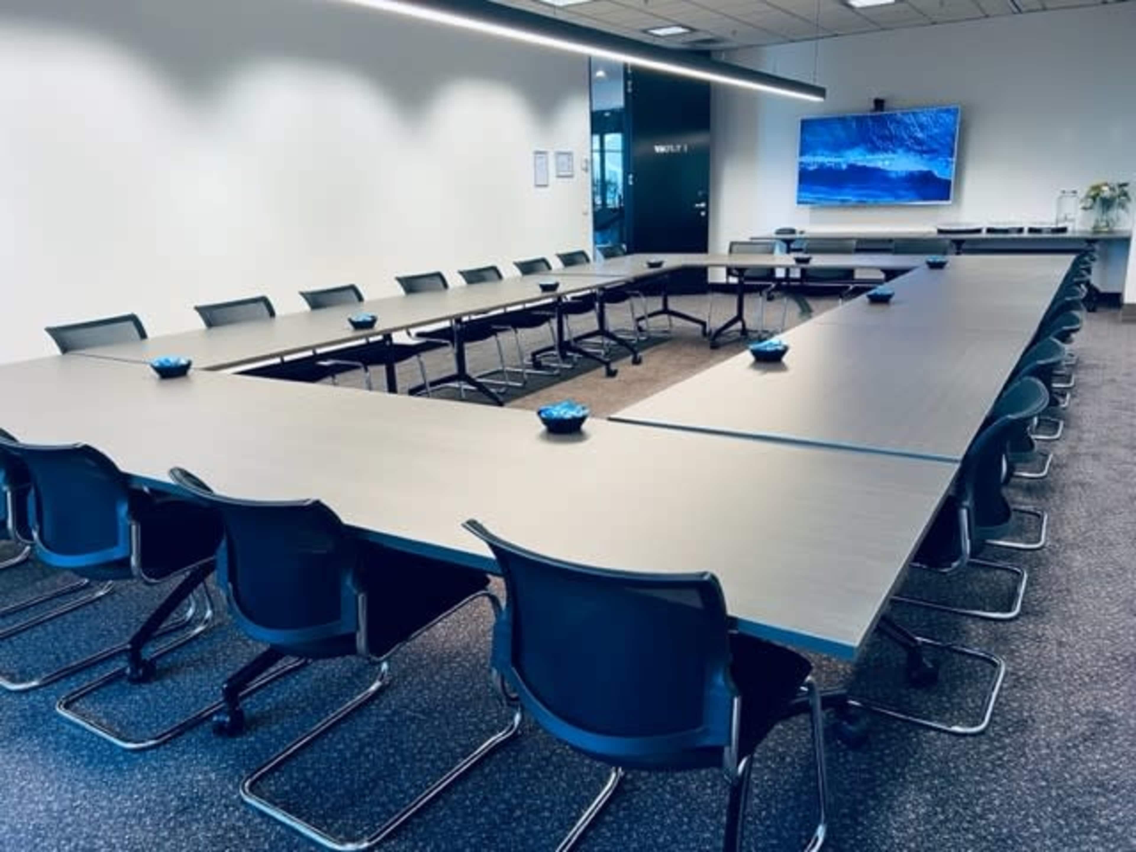 The image shows a modern conference room with a large U-shaped table arrangement and a mounted screen at one end.