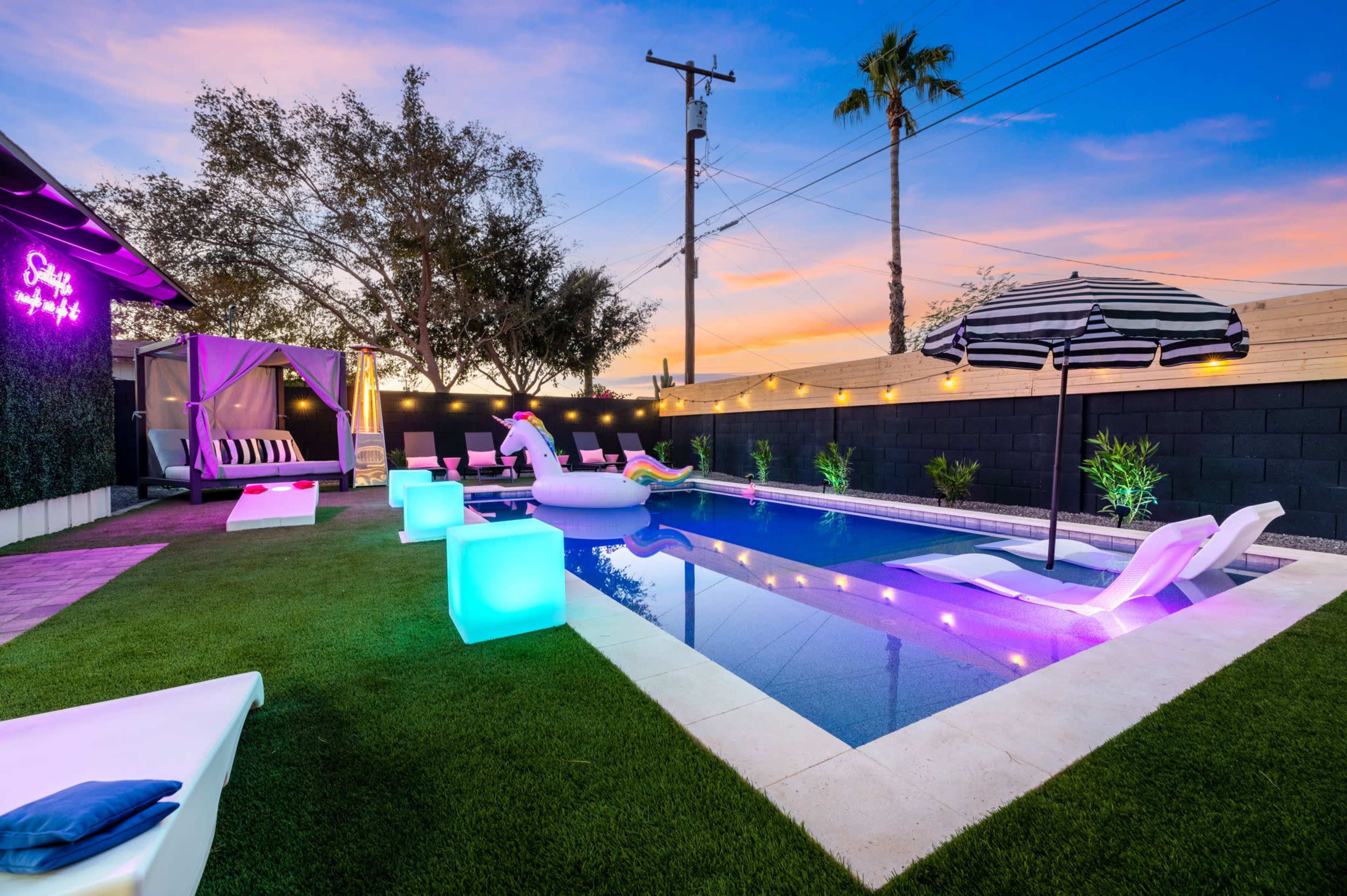 A backyard pool area with colorful lounge chairs, a large inflatable unicorn, and ambient lighting set against a sunset sky.