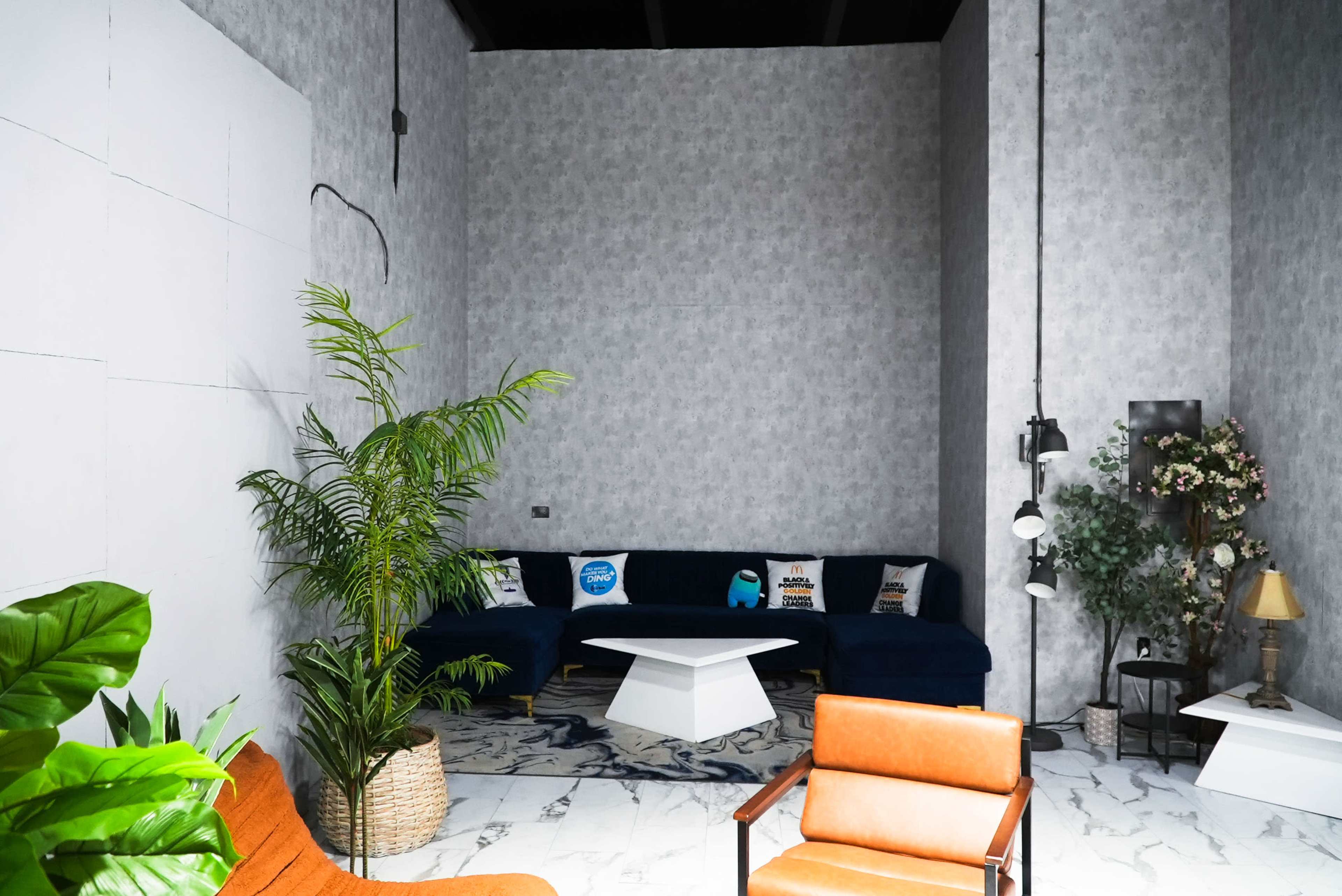 The image shows a modern, minimalist interior with a blue sofa, an orange chair, plants, and decorative items against a patterned gray wall.