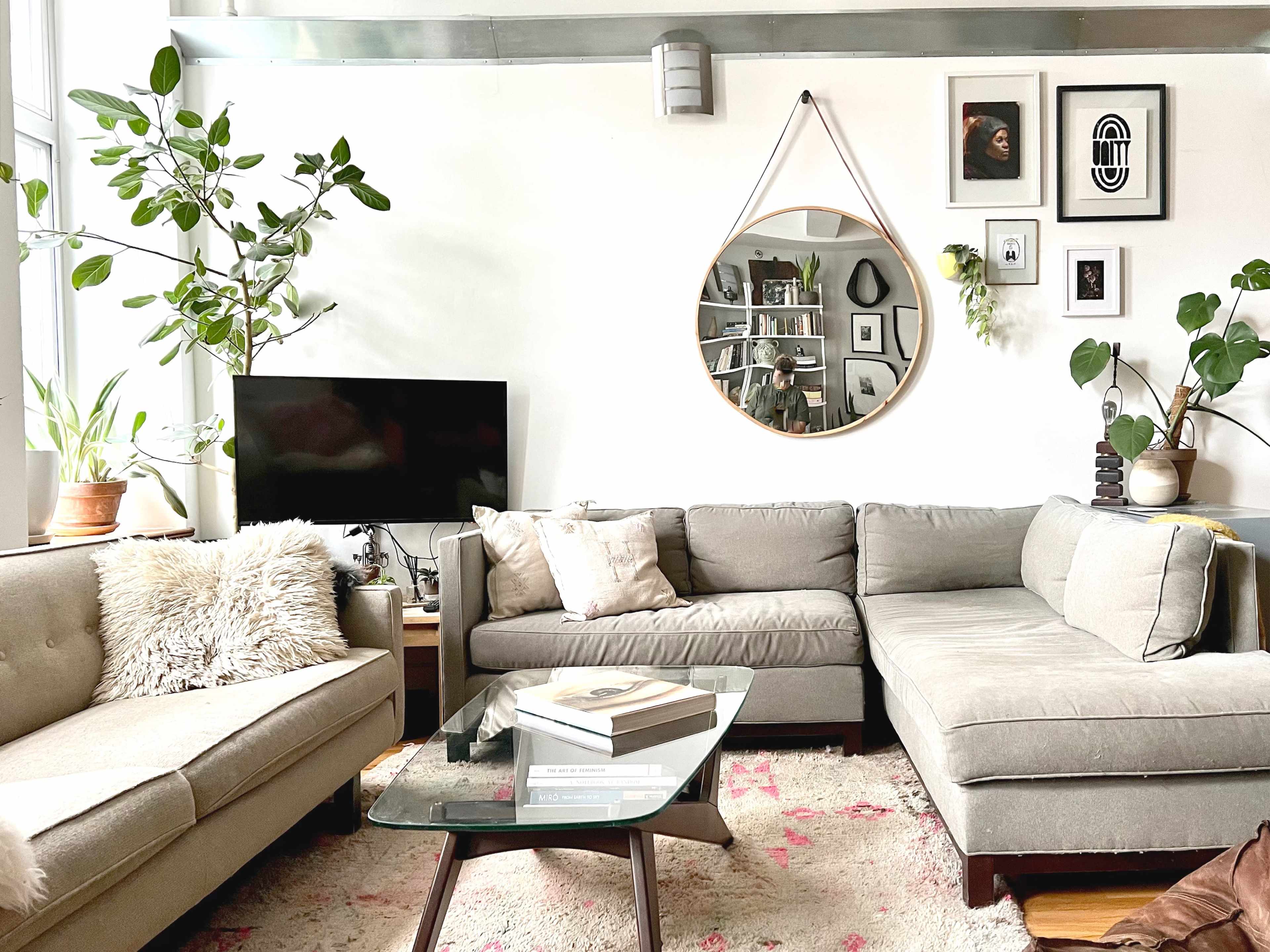 A bright living room features a light gray sectional sofa, a glass coffee table on a patterned rug, and a large round mirror hanging on the wall, surrounded by plants and framed artwork.
