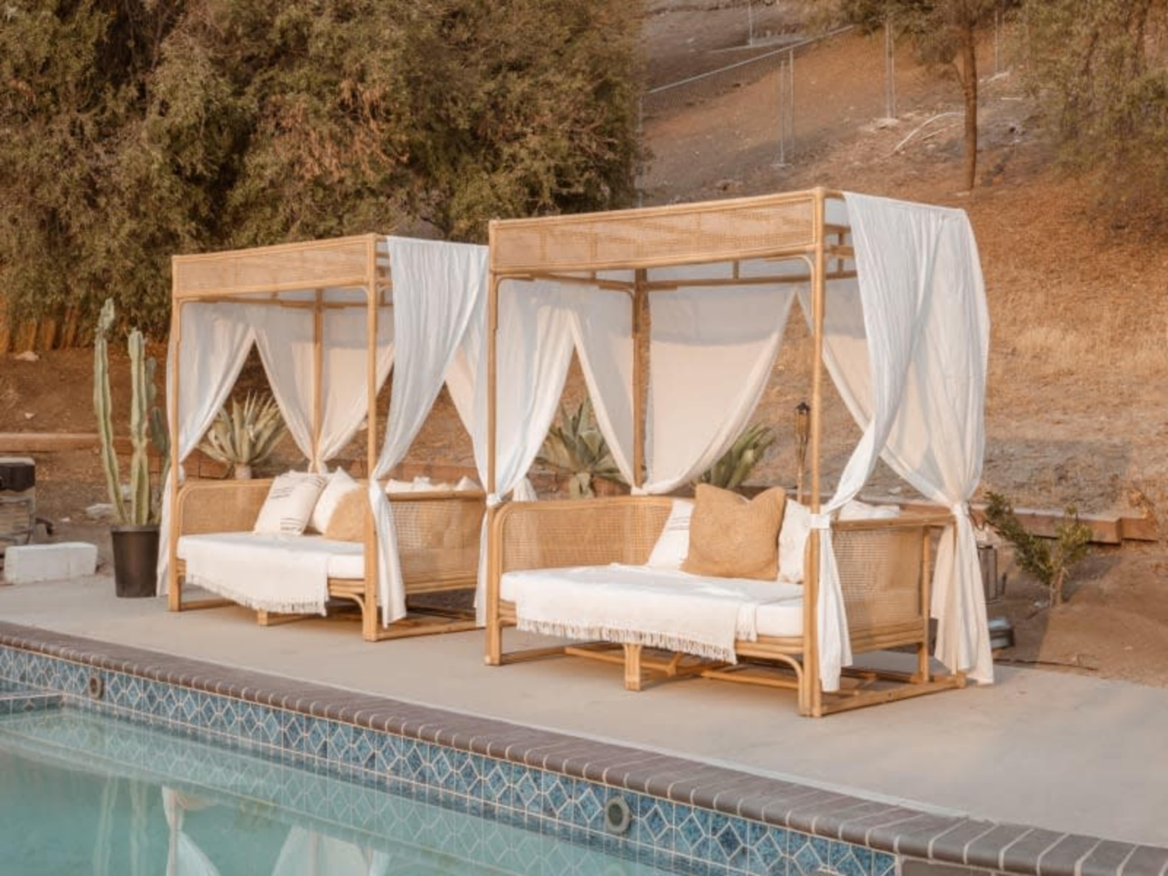 Two canopy lounge chairs with white drapes sit by a swimming pool surrounded by greenery.