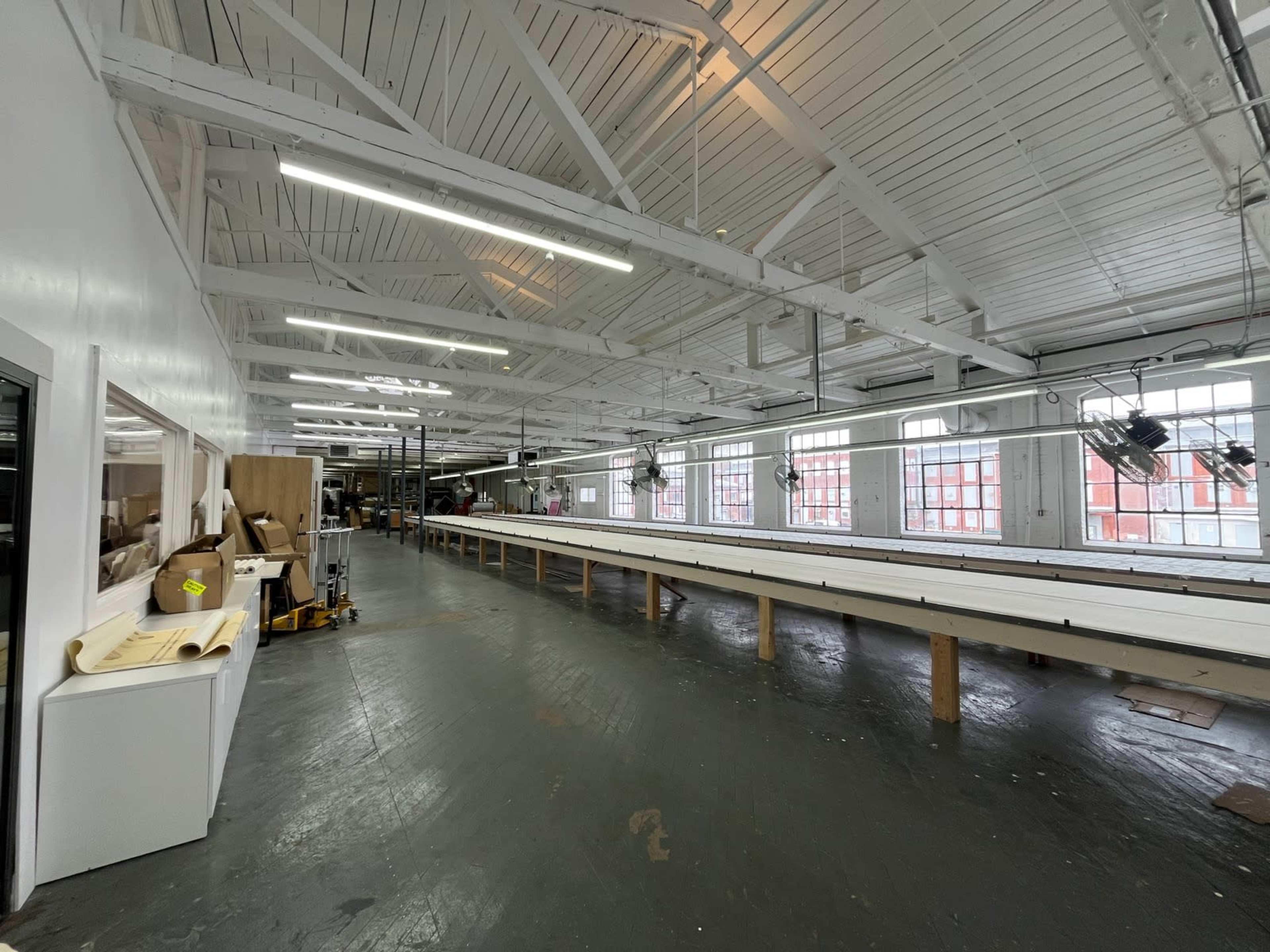 The image shows a spacious industrial workspace with large windows, wooden beams, and a long table running along one side.