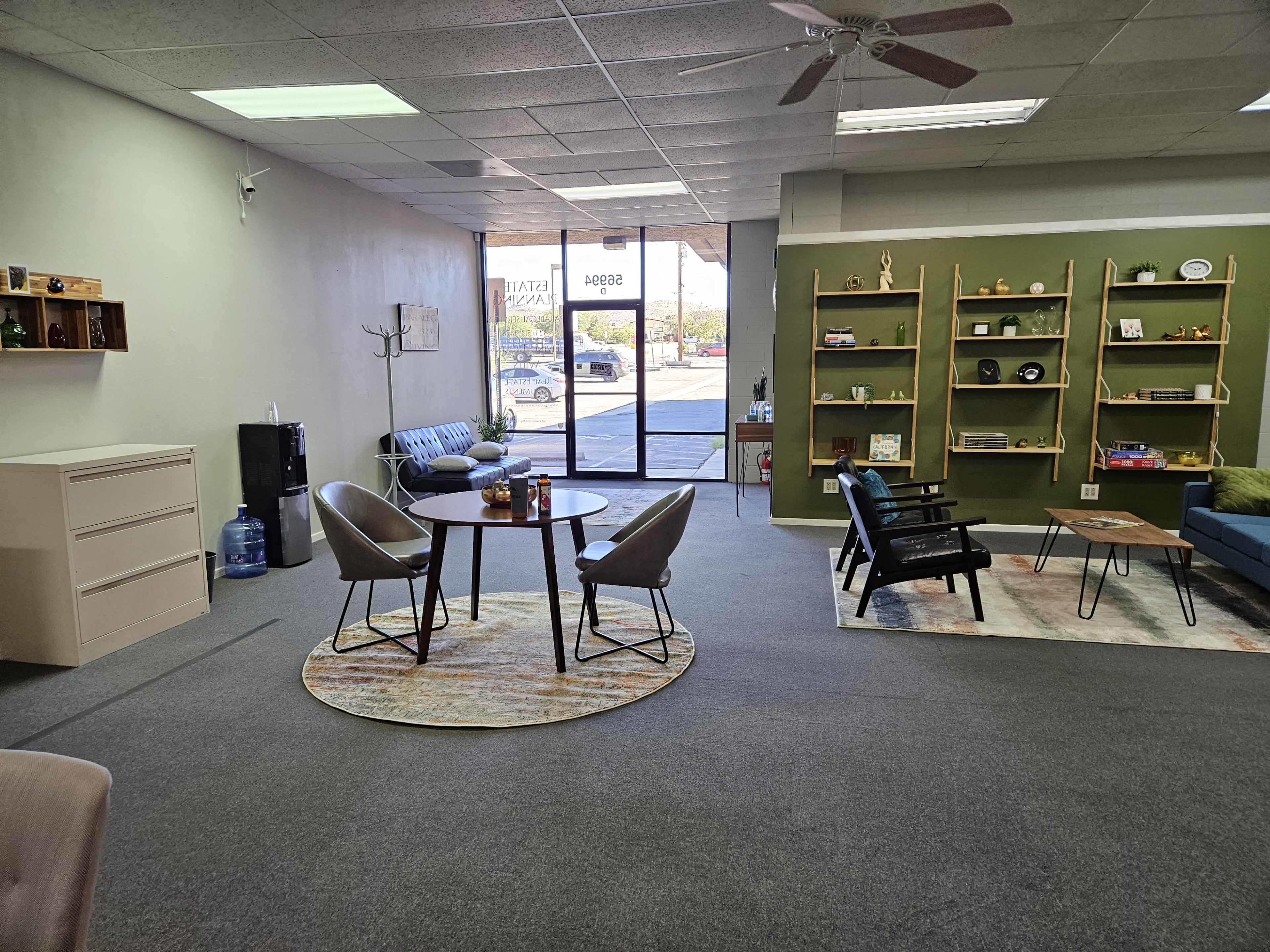 An interior space featuring a circular table with four chairs, a shelving unit against a green wall, and a seating area with a couch and chairs near large windows.